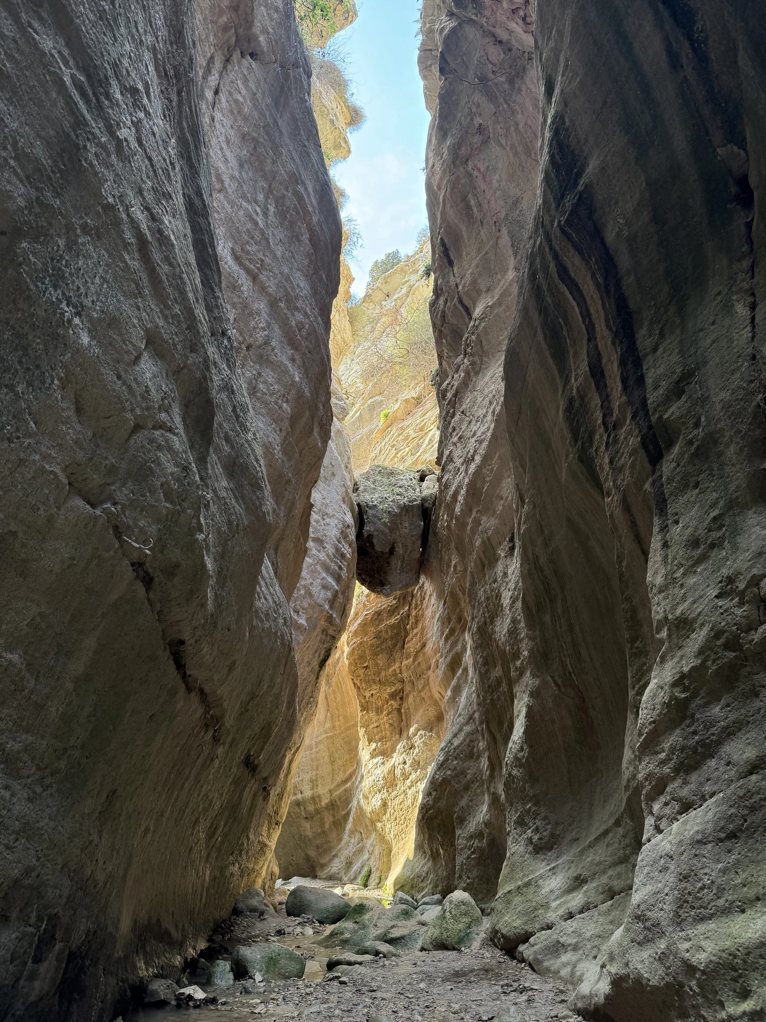 A tall, narrow canyon with steep rock walls and a big boulder suspended between the walls, with sunlight shining through the top opening.