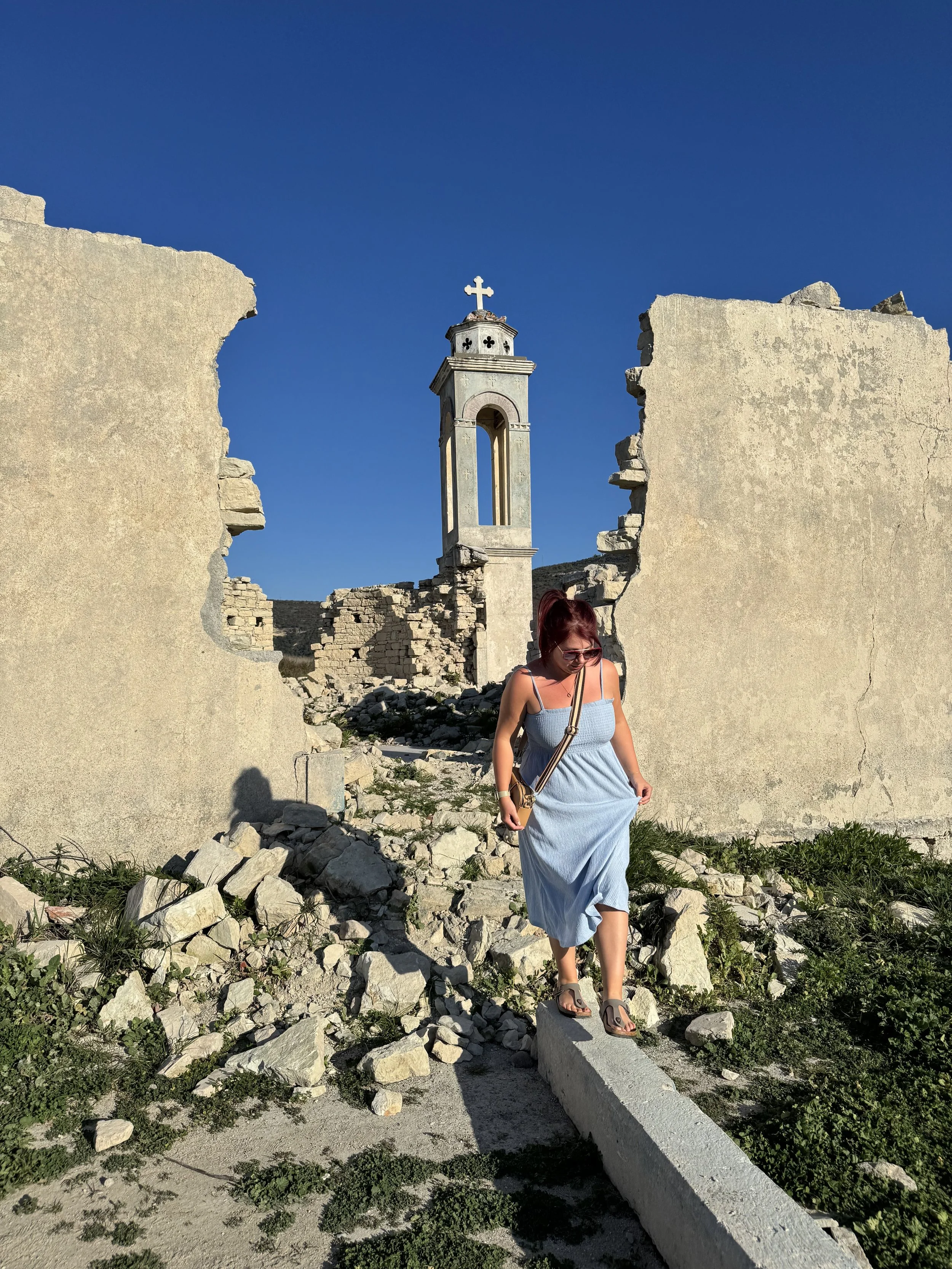 A woman in a blue dress and sandals walking along a narrow concrete path through ruins of a stone wall, with a small church or chapel with a bell tower and cross in the background, under a clear blue sky.