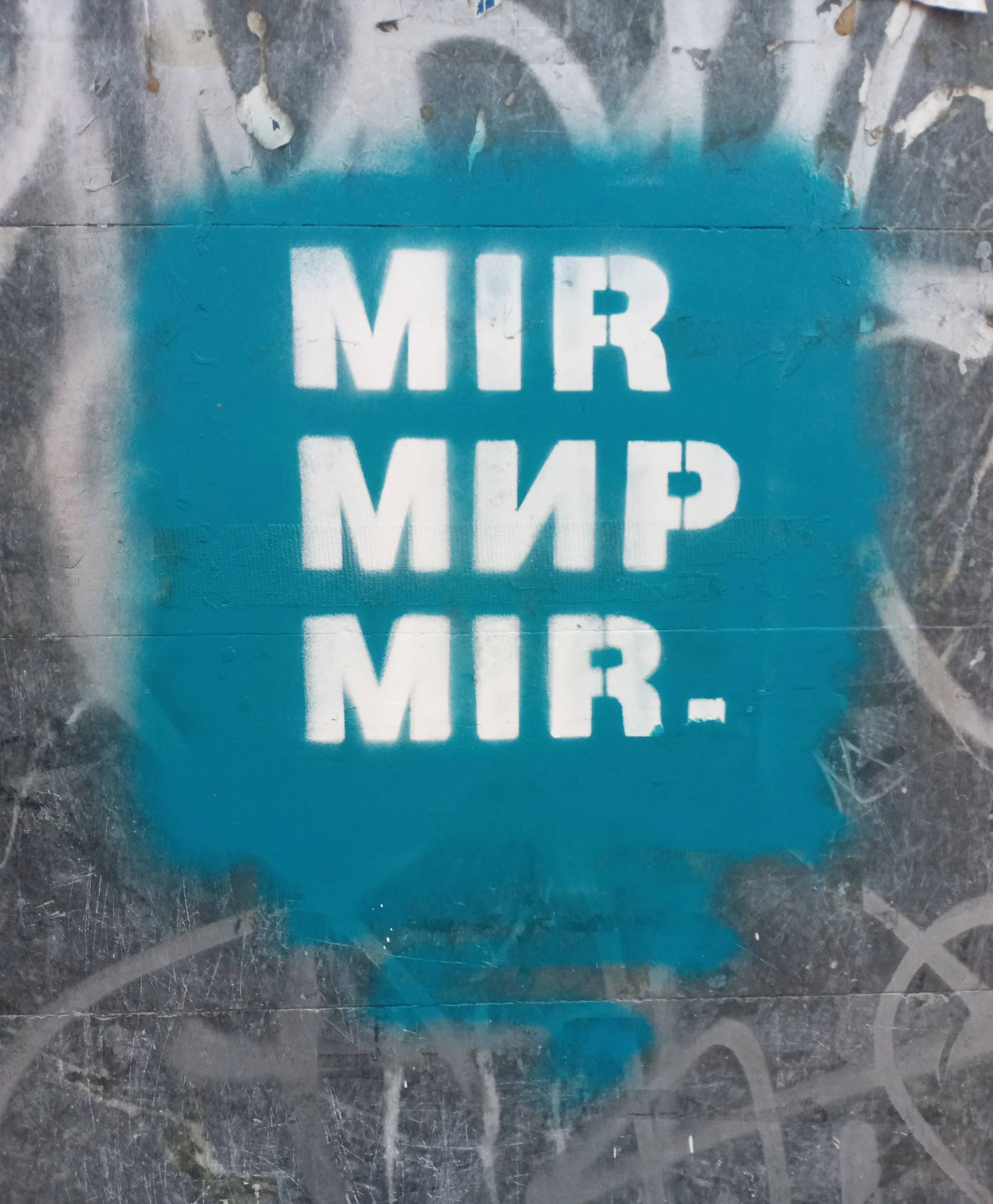 Stencil graffiti on a worn, gray wall reading “MIR MIR MIR.” in bold white letters, repeated three times over a turquoise spray-painted rectangle, with surrounding layers of faded tags and scratches.