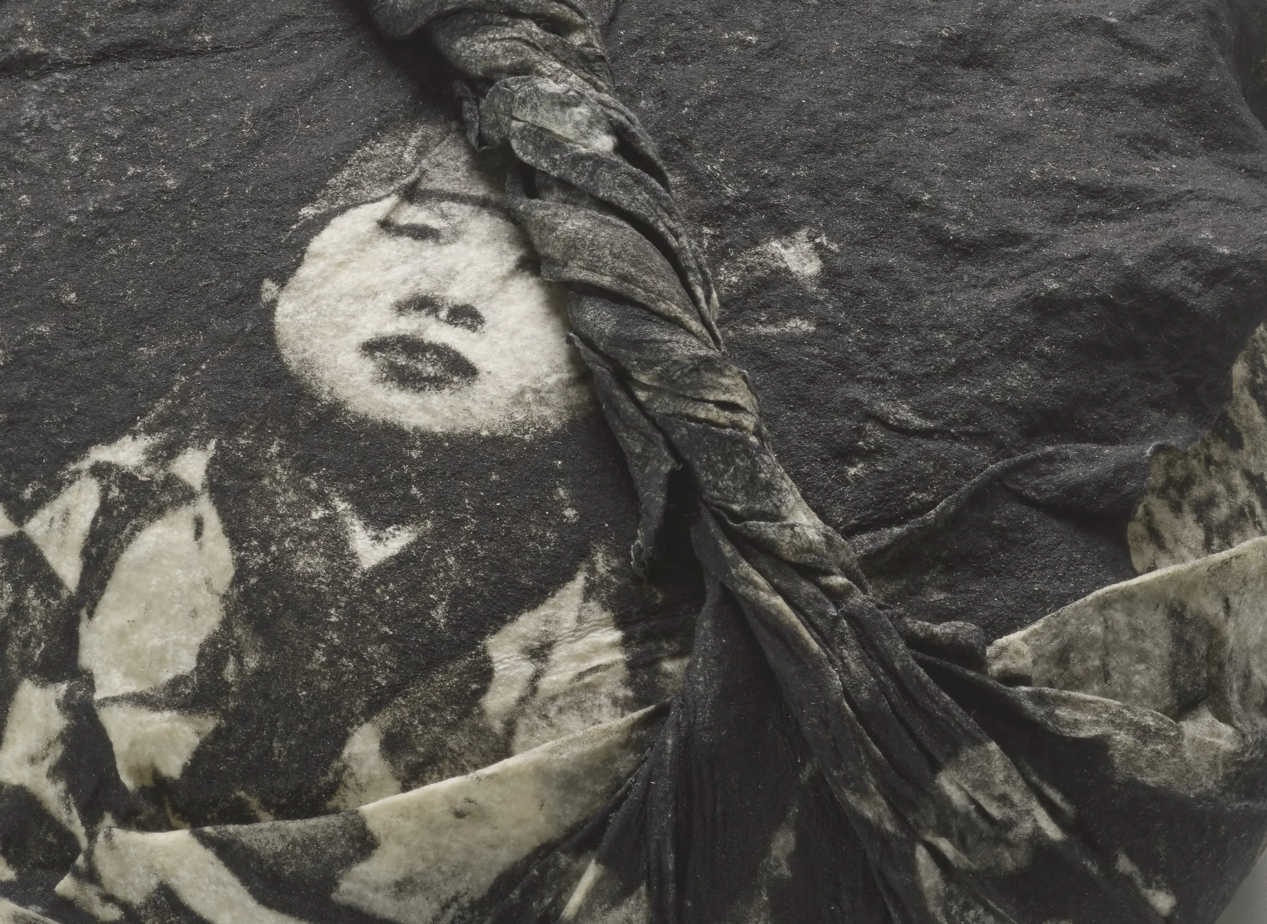 Weathered black-and-white textile showing a woman's face with eyes closed, beside a thick braided twist of the same material.