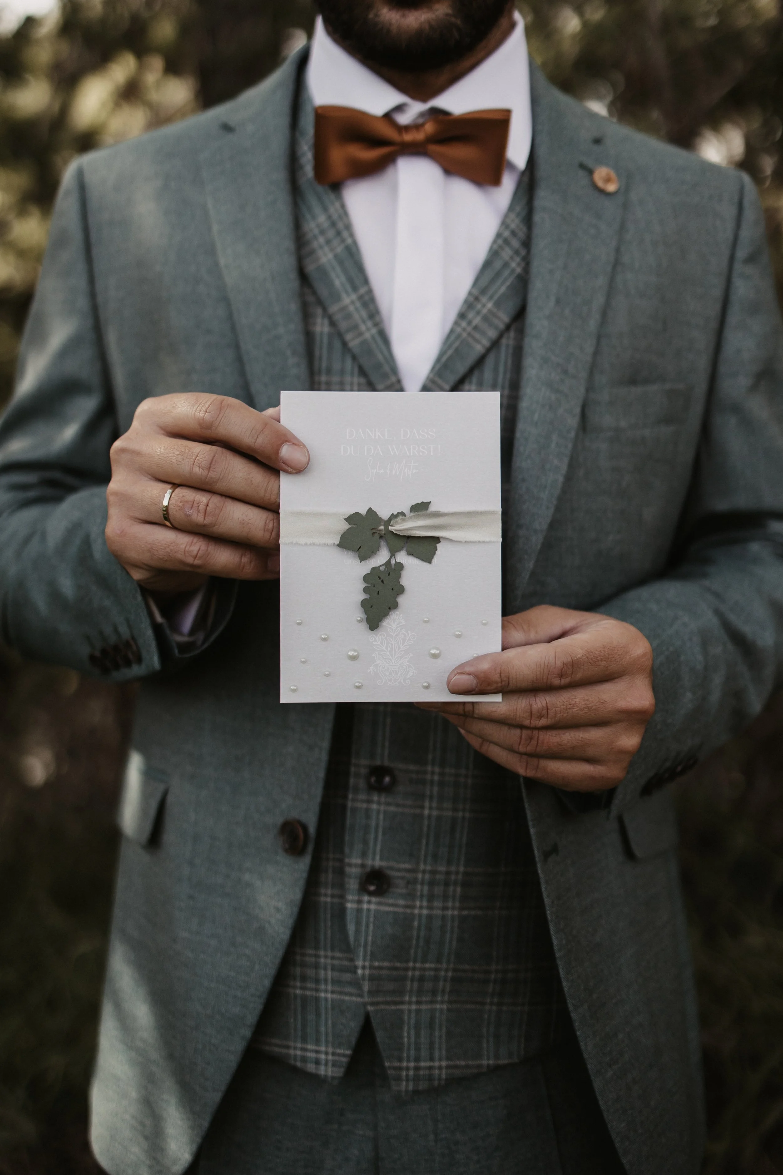 Ein Mann in formeller Kleidung hält eine Grußkarte mit Weinreben-Motiv. Er trägt einen grauen Anzug, Fliege und ein Hemd mit Karo-Muster, die Karte ist zur Kamera gerichtet.