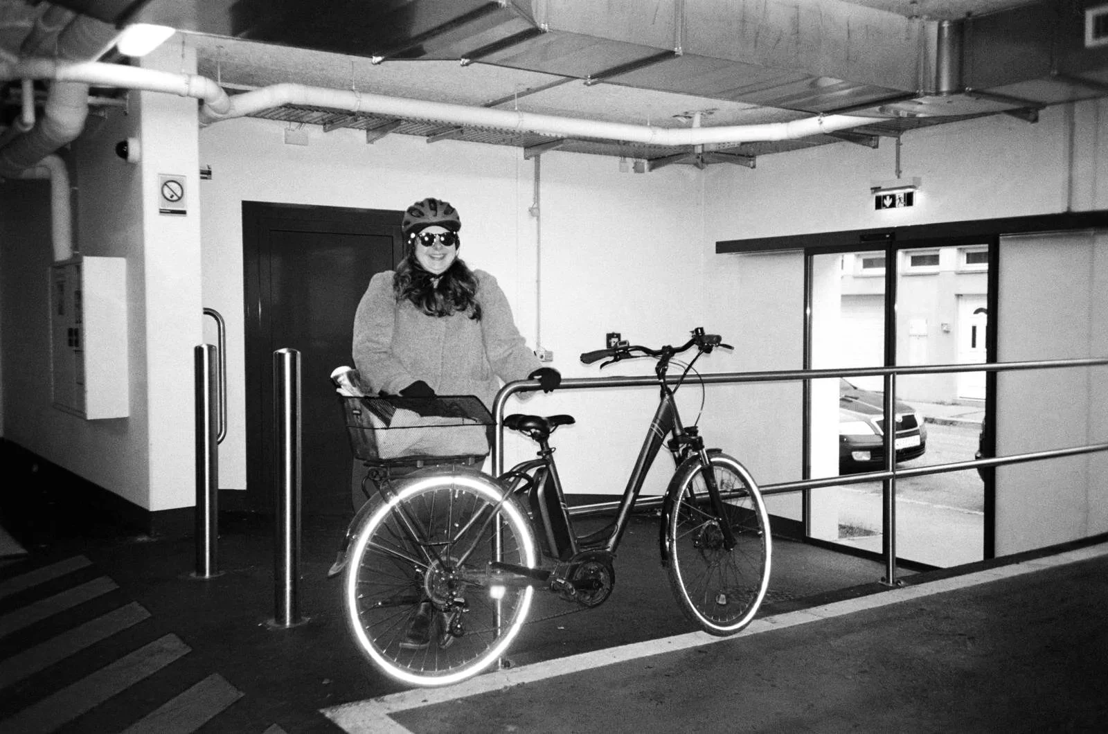 Eine Frau mit Fahrrad in einer Tiefgarage, trägt einen Helm und Sonnenbrille, sie ist lachend und hat eine Tasche im Fahrradkorb.