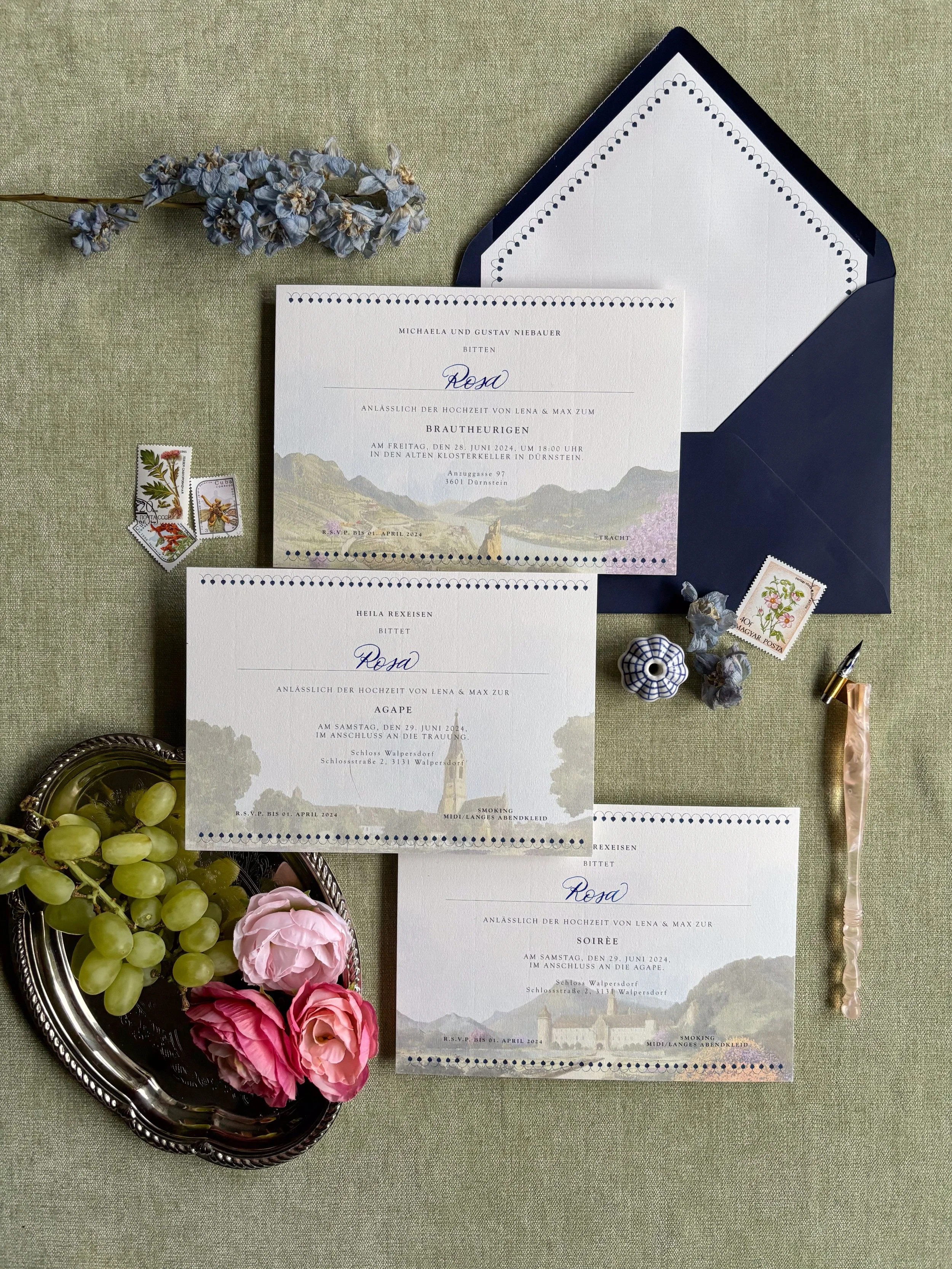 Ein Einladungssatz für eine Hochzeit mit Briefumschlag, Blumendekoration, Weintrauben, einer Siegelkapsel, Briefmarken, einer Schreibfeder und Hochzeitskarten auf einem grünen Stofftuch.