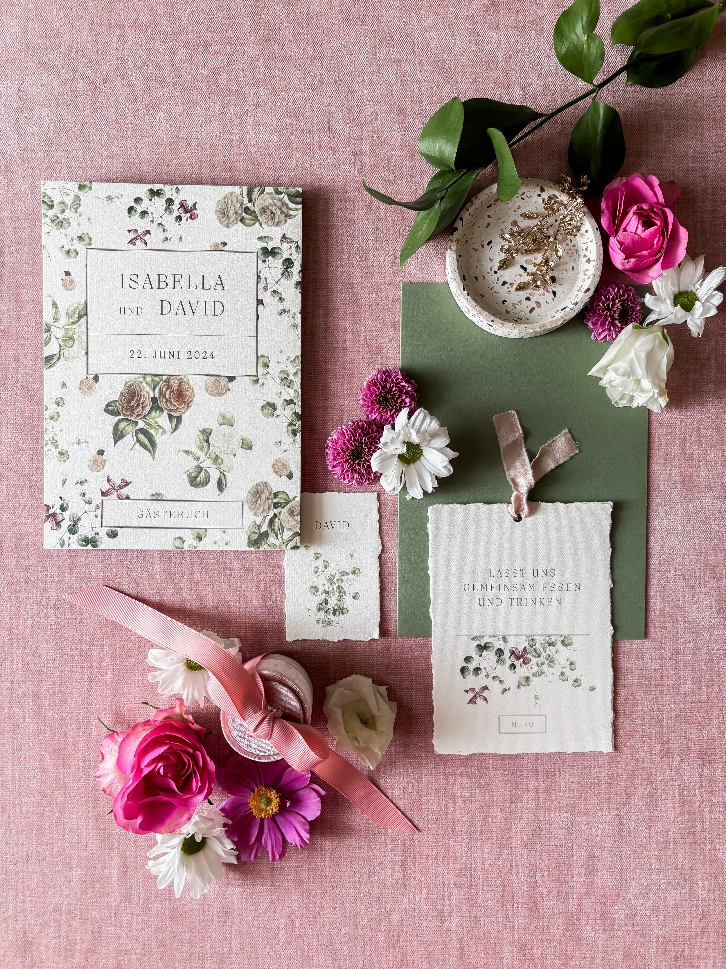 Hochzeitseinladung mit Blumen und Dekoration auf rosa Stoff, inkl. Einladungskarte, Gästebuch, Menu-Karte, kleine Zettel, Blumen, Kerze, und einer Pflanze.