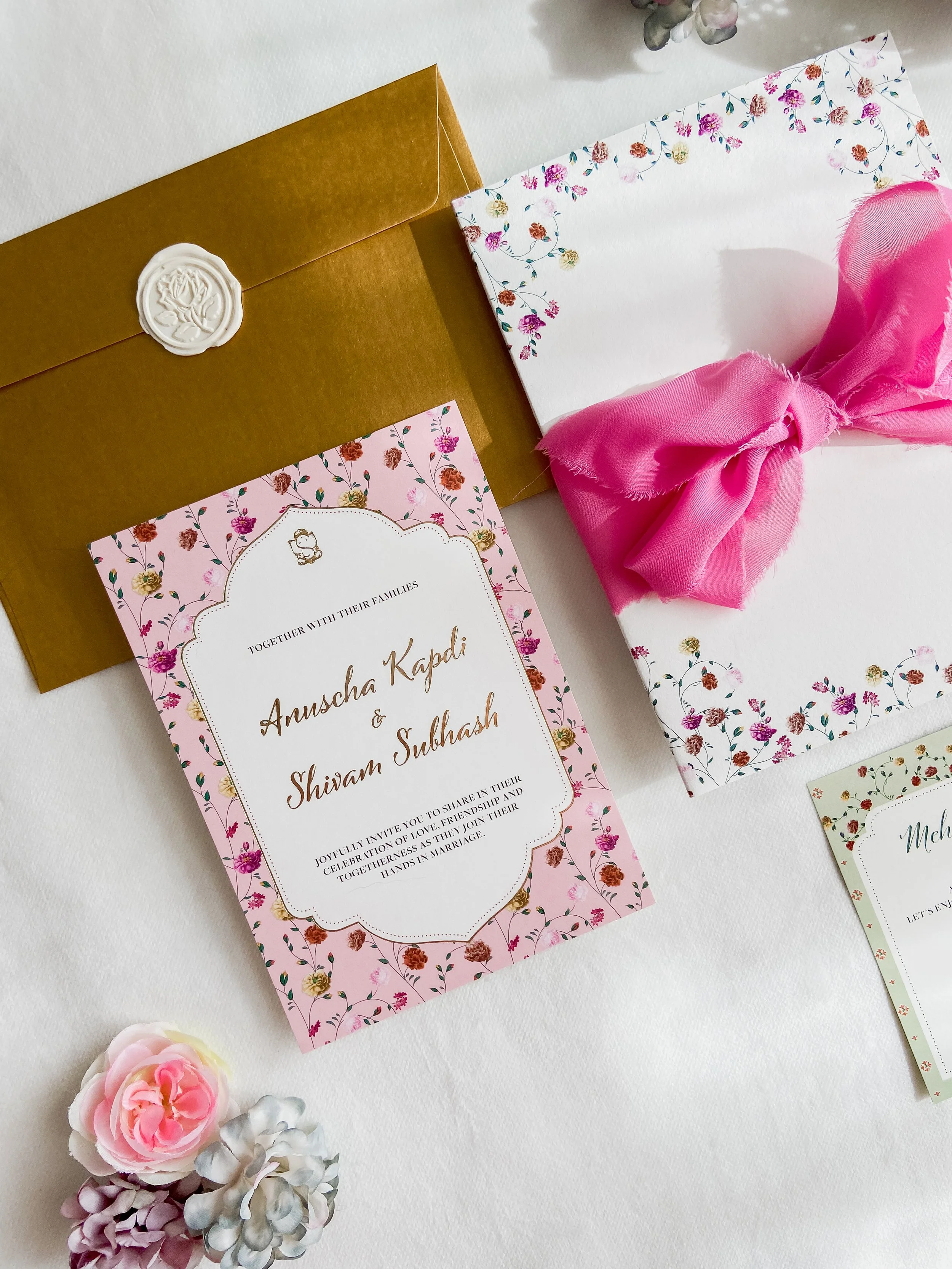 Hochzeitseinladung, goldener Umschlag mit weißem Siegel, rosa und weiße Blumenkarte mit goldener Schrift, Geschenkumschlag mit rosa Schleife, Blumen im Vordergrund