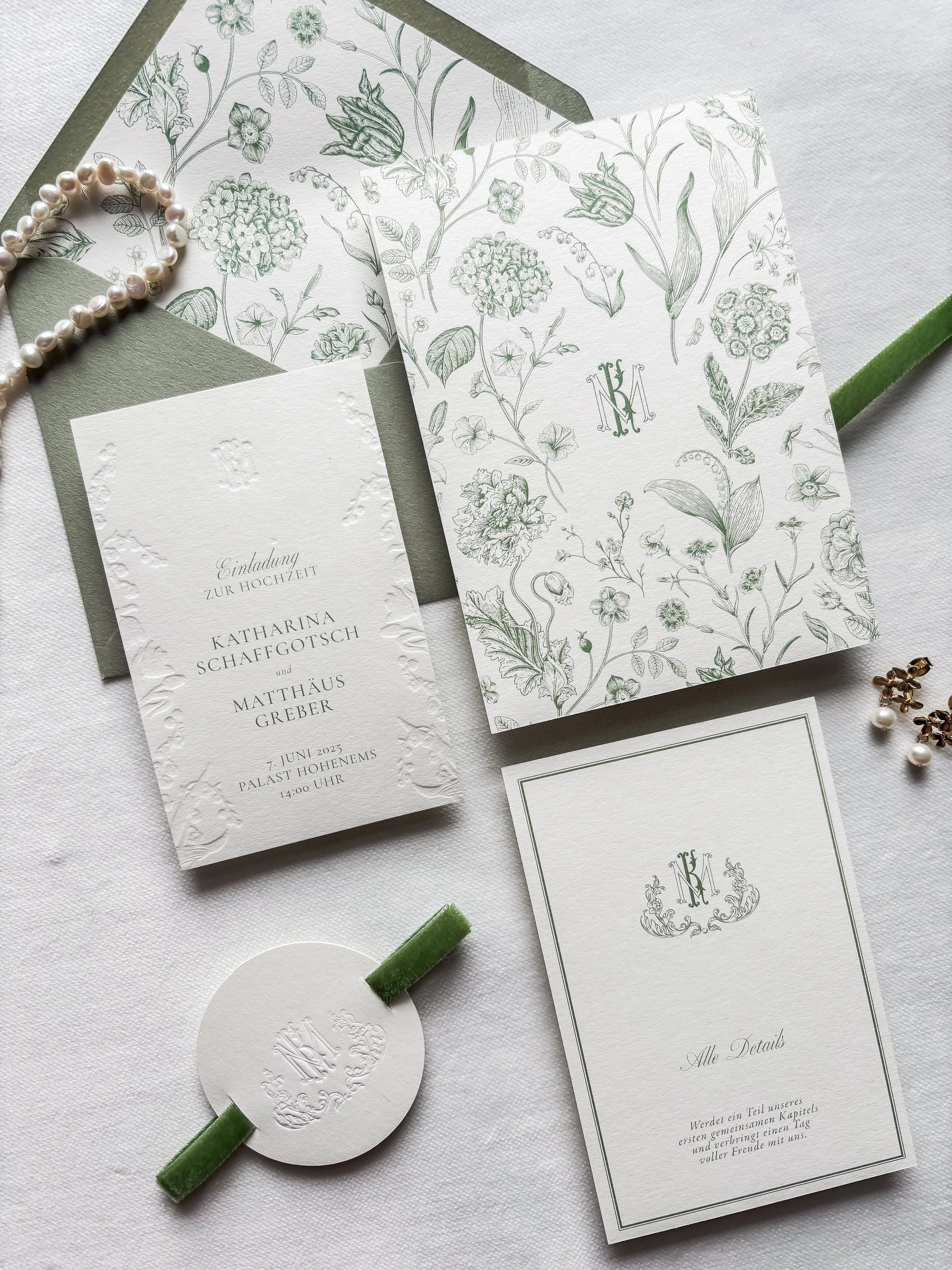 Eine Hochzeitskarte mit floralem Muster, einer Einladungskarte, einem grünen Seidenband, einem kurzen weißen Kartenetikett mit Monogramm, und einem weiteren Faltblatt mit Monogramm und Text, alles auf weißem Stoffuntergrund