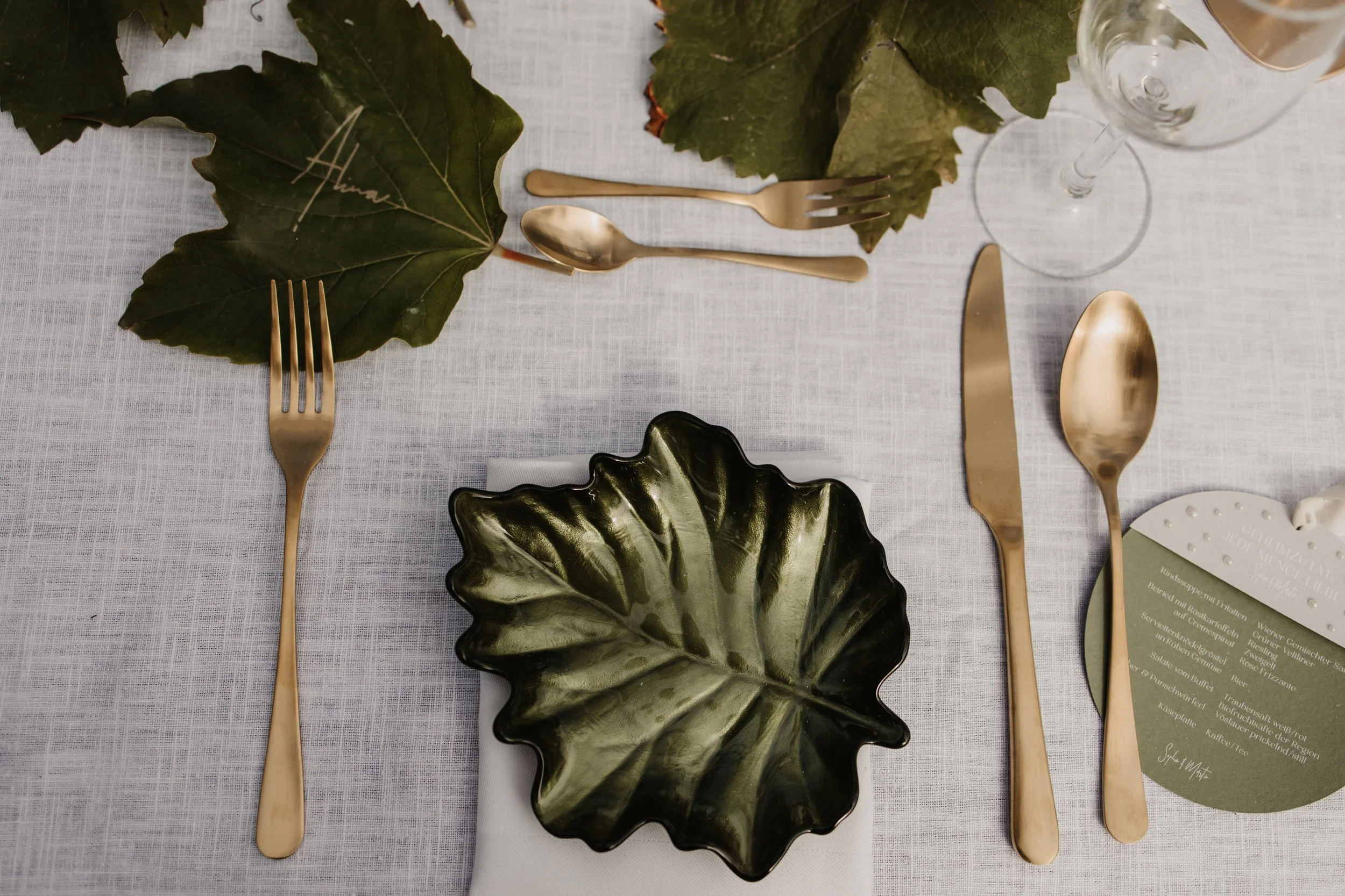 Hochzeitstafel mit blattförmigem Teller, goldenem Besteck als Highlight und graviertem Weinblatt Namenskärtchen