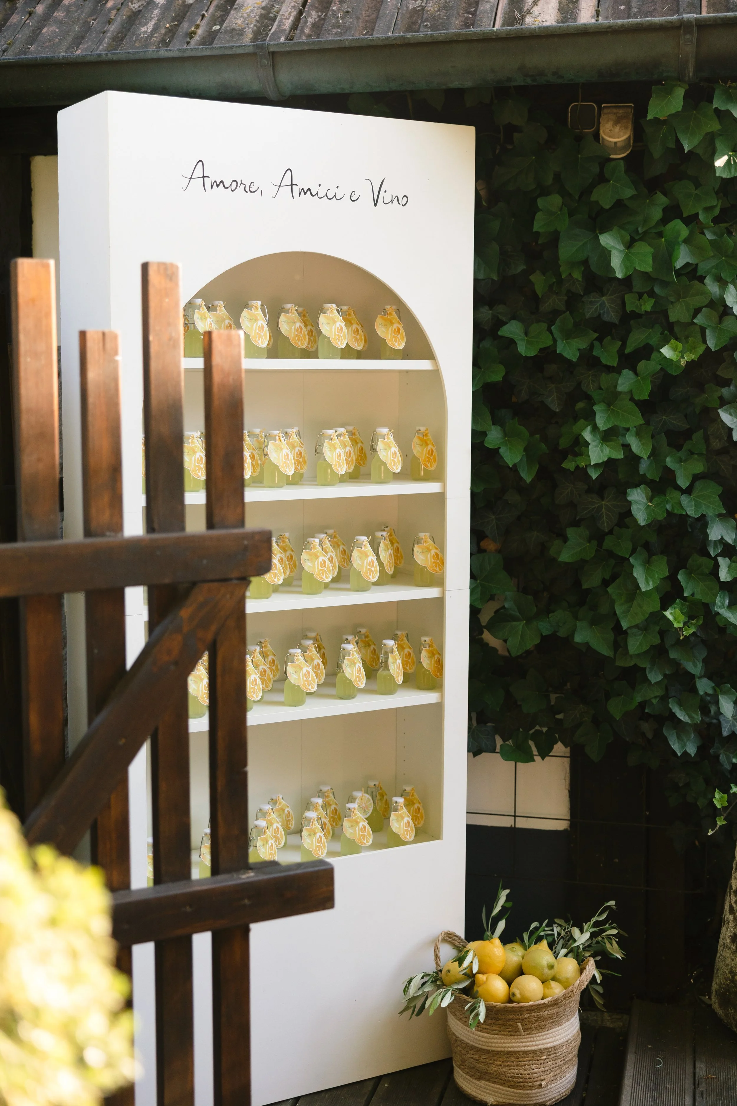 Weiße Verkaufsständer mit Zitronenlimonade-Flaschen, beschriftet mit 'Amore, Amici e Vino', im Garten mit grünem Laub im Hintergrund und einer Korb voller Zitronen davor.