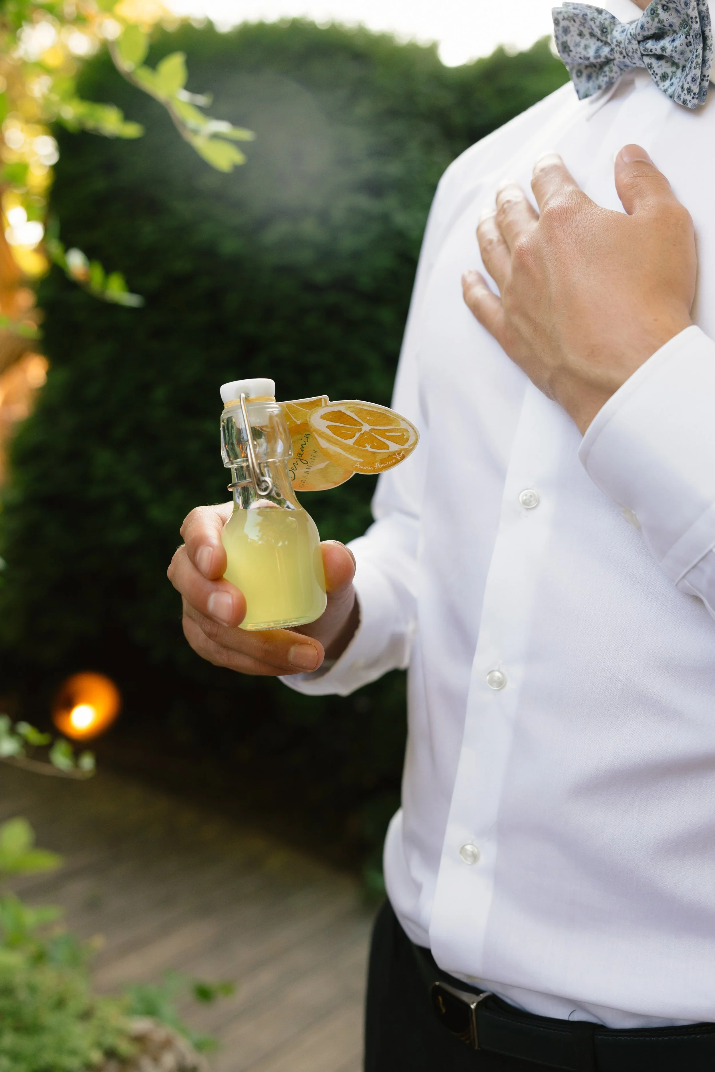 Mann in weißem Hemd mit Fliege hält eine kleine Flasche Zitronenlimonade, während er seine Hand auf die Brust legt, im Außenbereich bei Tageslicht.