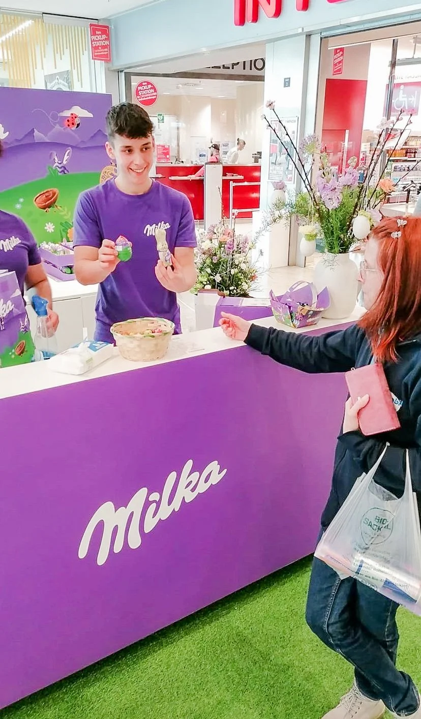 Eine junge Frau kauft an einem Milka-Schalter Schokoladenprodukte im Supermarkt, während ein männlicher Verkäufer ihr ein Produkt zeigt. Der Schalter ist lila mit dem Milka-Logo und dem Hintergrund ist bunte Dekoration mit Blumen.