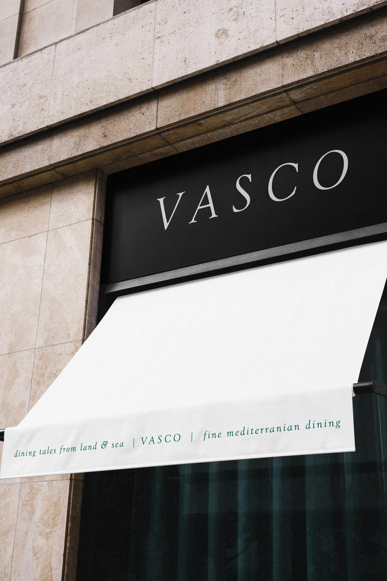 Das Bild zeigt ein schwarzes Schild mit dem weißen Schriftzug 'VASCO'. Darunter hängt ein weißes Markisentuch mit grüner Schrift, auf dem steht 'dining tales from land & sea | VASCO | fine mediterranean dining'. Der Hintergrund besteht aus einer Steinwand.