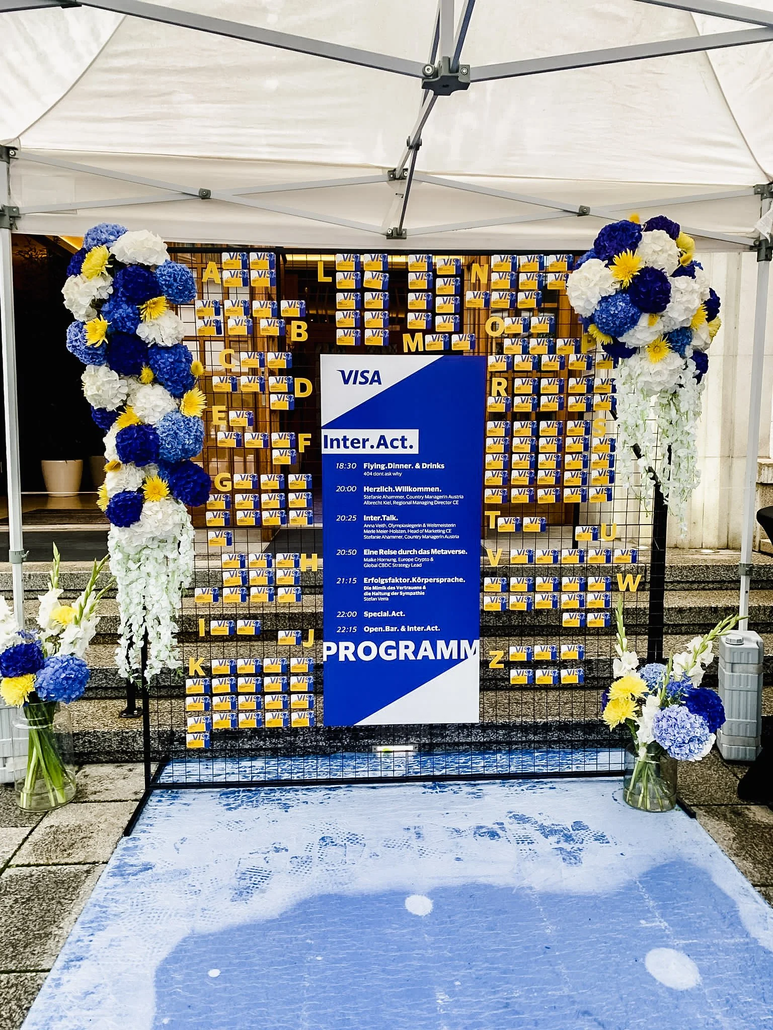 Ein Veranstaltungsschild mit Programmübersicht, umgeben von blauen, weißen und gelben Blumen, mit VISA-Logos im Hintergrund und einer weißen Plane darüber.