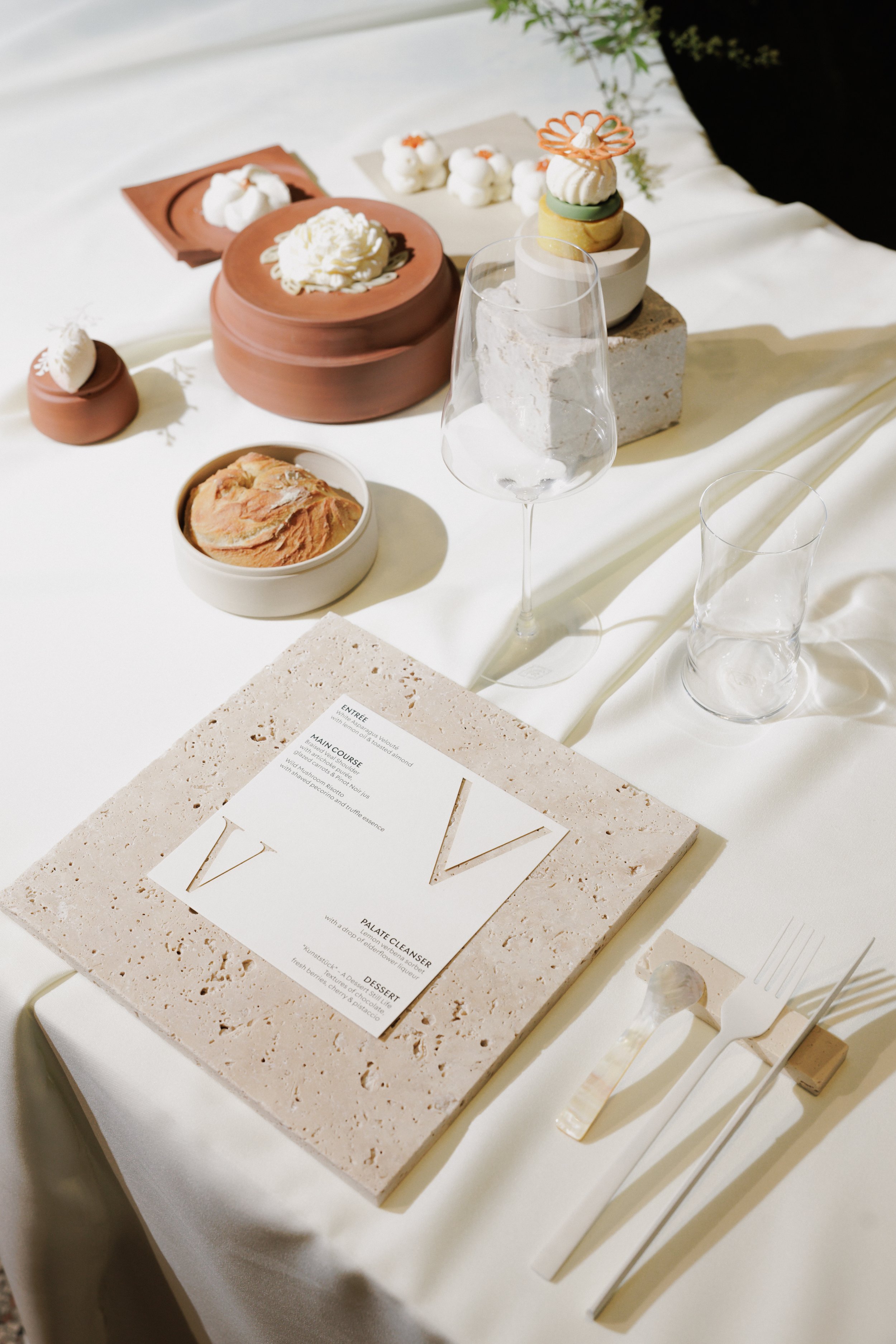 Eleganter gedeckter Esstisch mit Speisekarte, Weingläsern, Besteck, Keramikdekoration und Süßspeisen, einschließlich Schlagsahne, Gebäck und kleinen Torten, mit hellen, neutralen Farben.