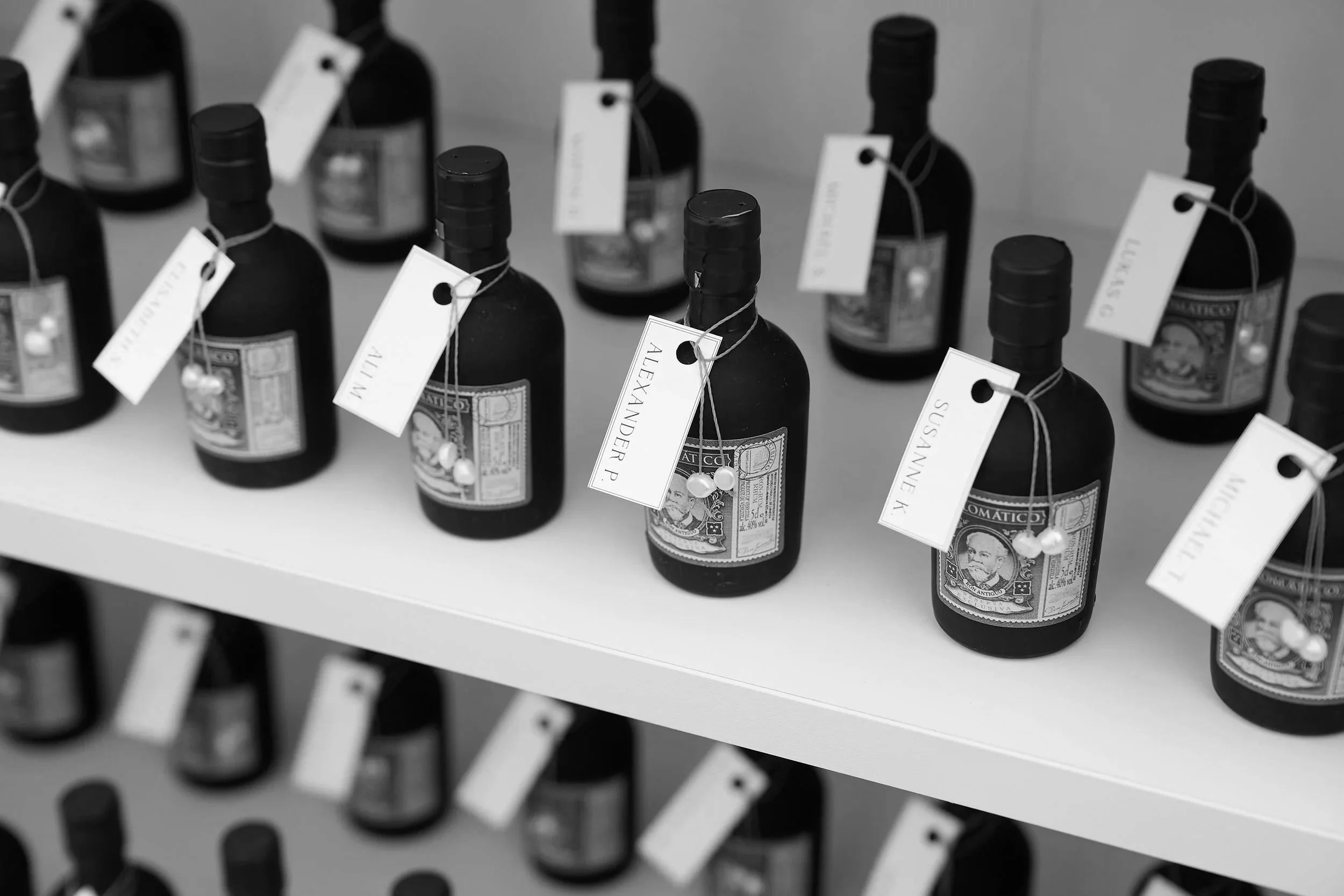 Schwarz-weiße Aufnahme von mehreren Weinflaschen auf einer weißen Regalfläche, jede Flasche ist mit einem weißen Tag mit Name und Perlen verziert.