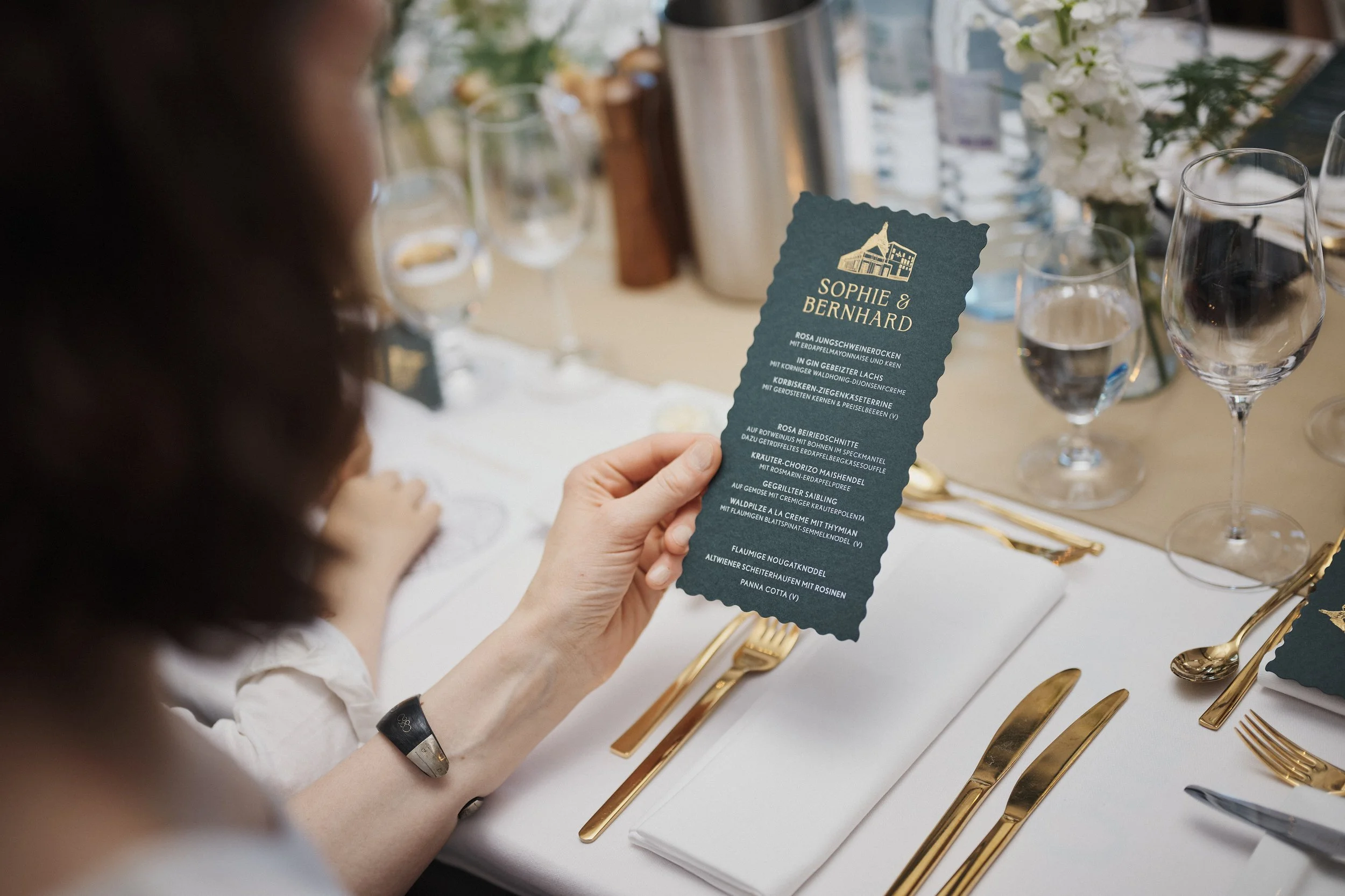Eine Frau hält eine Speisekarte bei einer eleganten Tischdekoration, bestehend aus goldenen Bestecken, Weingläsern, weißen Blumen und einem weißen Tischtuch.