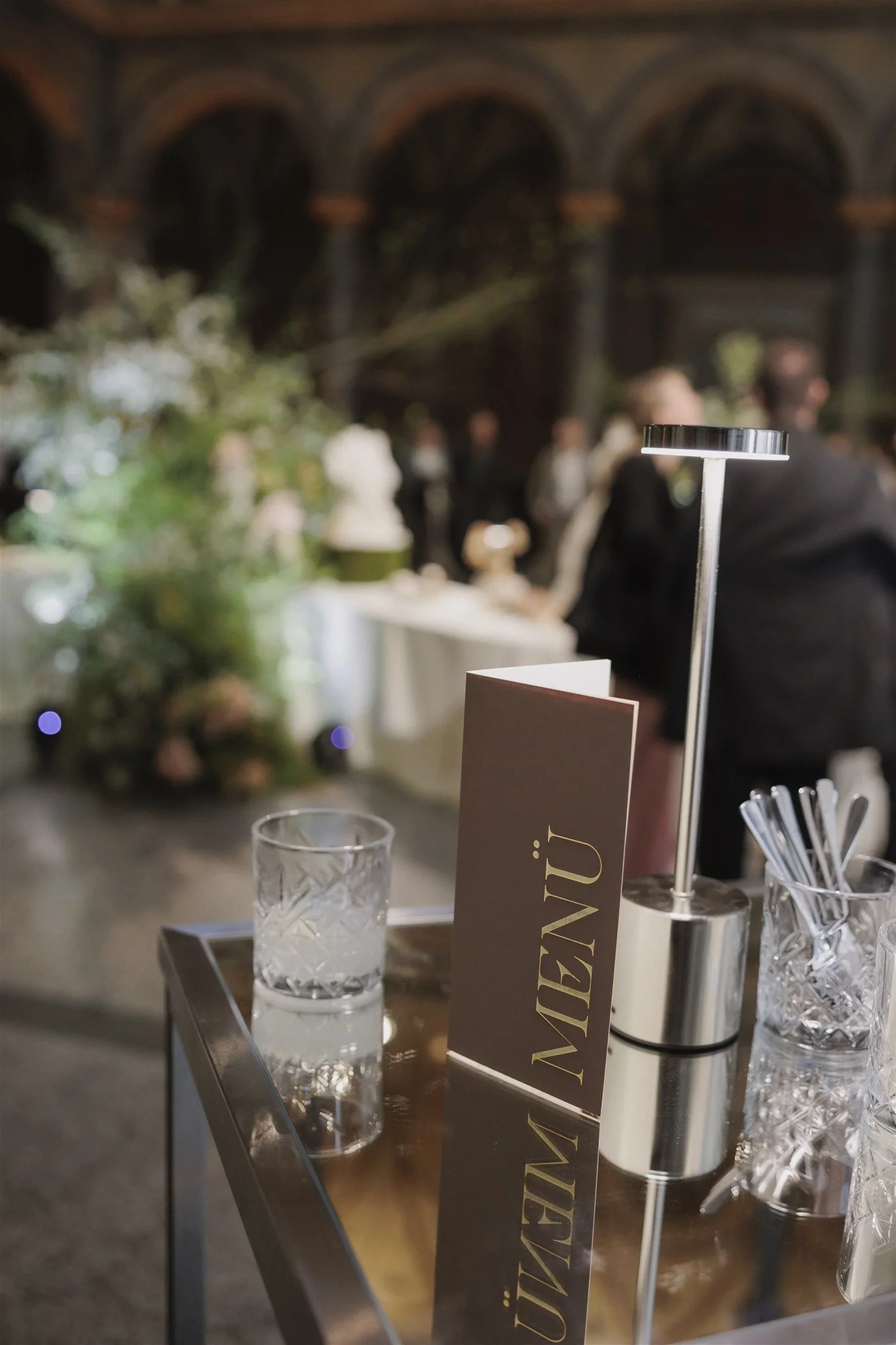 Ein eleganter Empfangstisch mit einem Willkommensschild, einem Glas Wasser, Strohhalmen und einem stilvollen Tischlampen in einem festlich dekorierten Veranstaltungsraum im Hintergrund.
