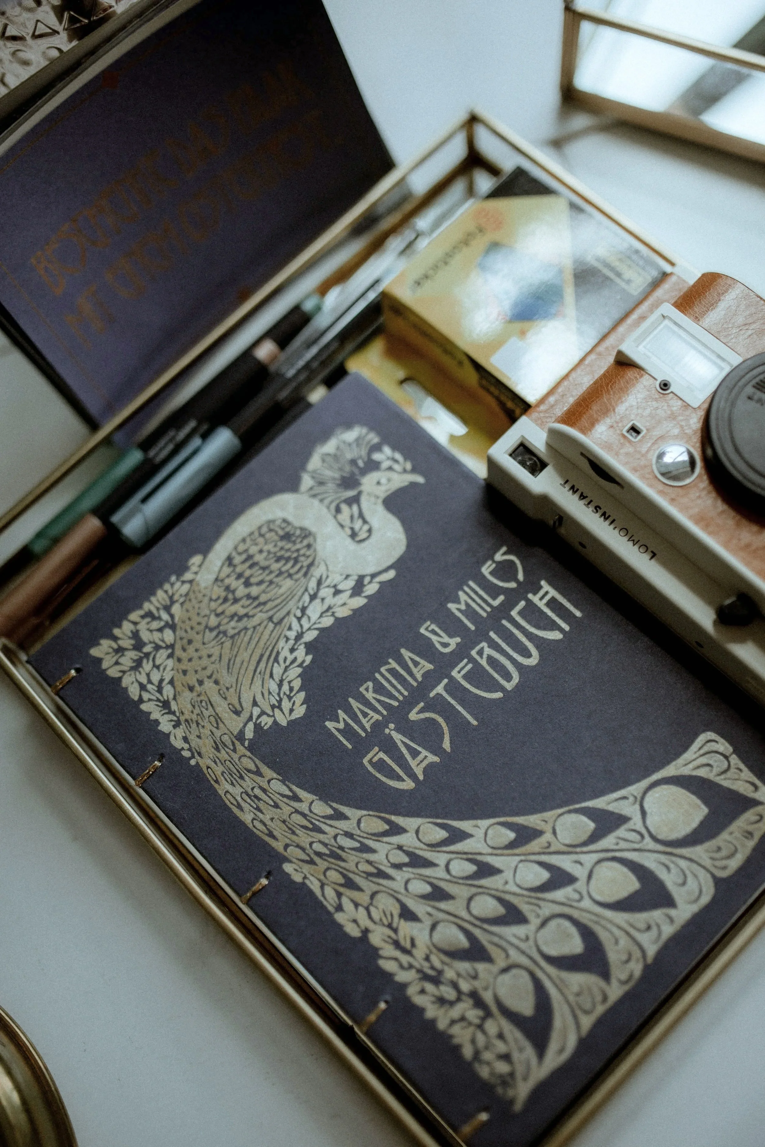 Buch mit goldener Illustration eines Pfauen auf einem schwarzen Hintergrund, mit den Namen Marina & Miles Ostertag auf dem Titel, in einem goldfarbenen Kartenetui mit anderen Schreibutensilien und einer Kamera.