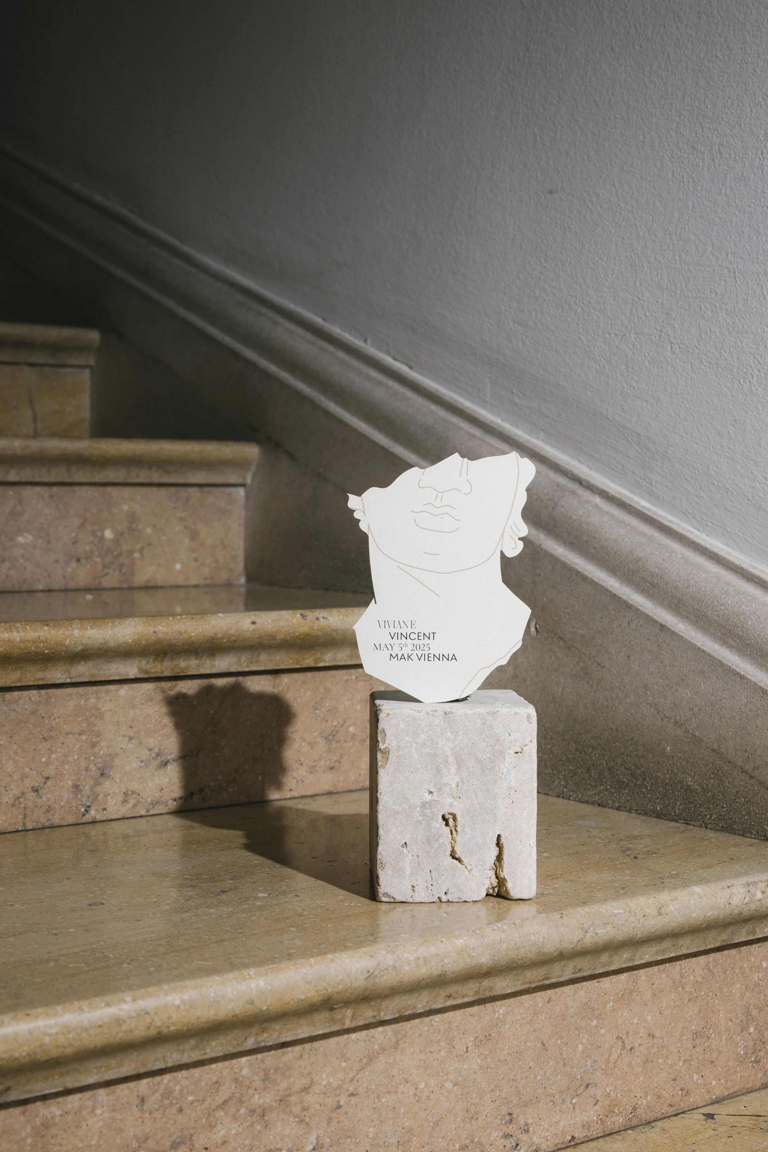 Ein Kunstwerk auf einem weißen Stein auf einer Treppe, das eine lineare Silhouette eines Gesichts und Text mit Namen, Datum und Ort zeigt.