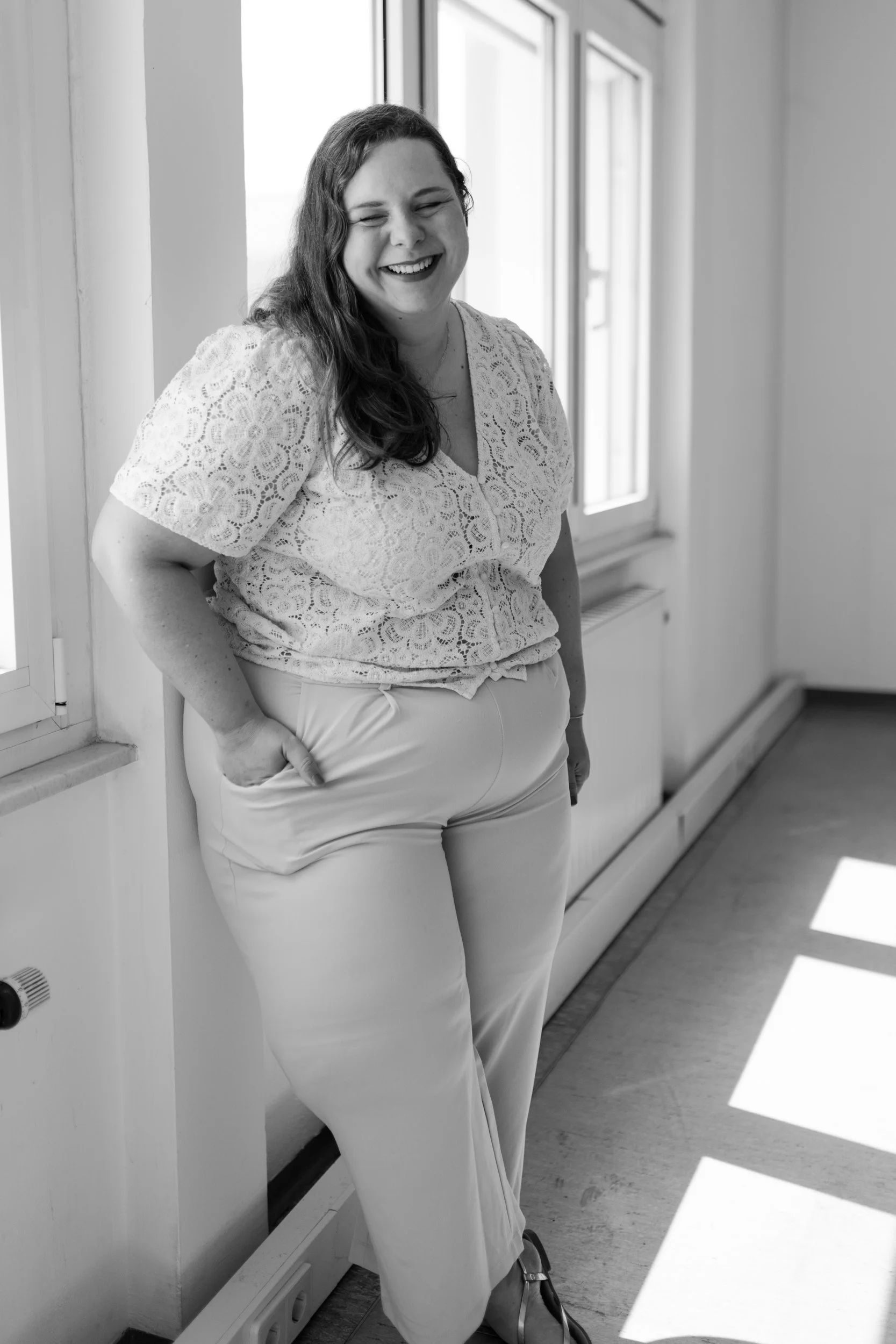 Eine lachende Frau lehnt an einer Fensterbank in einem Raum, schwarz-weiß Fotografie.