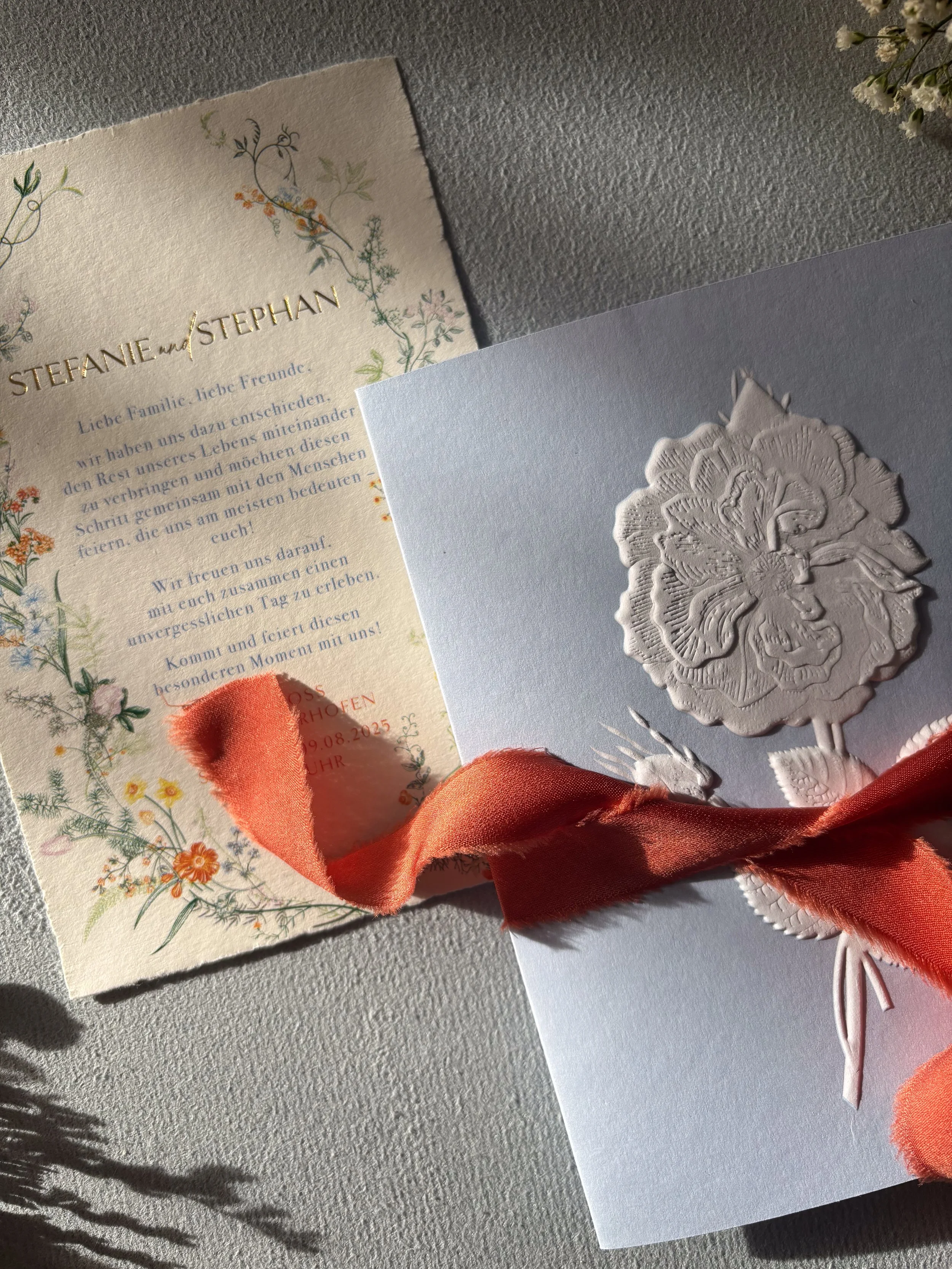 Ein Hochzeits-Einladung mit floralem Design, auf der Namen Stephanie und Stephan stehen, sowie eine weiße Karte mit floralem Relief und einem rosa Schleifenband darauf.