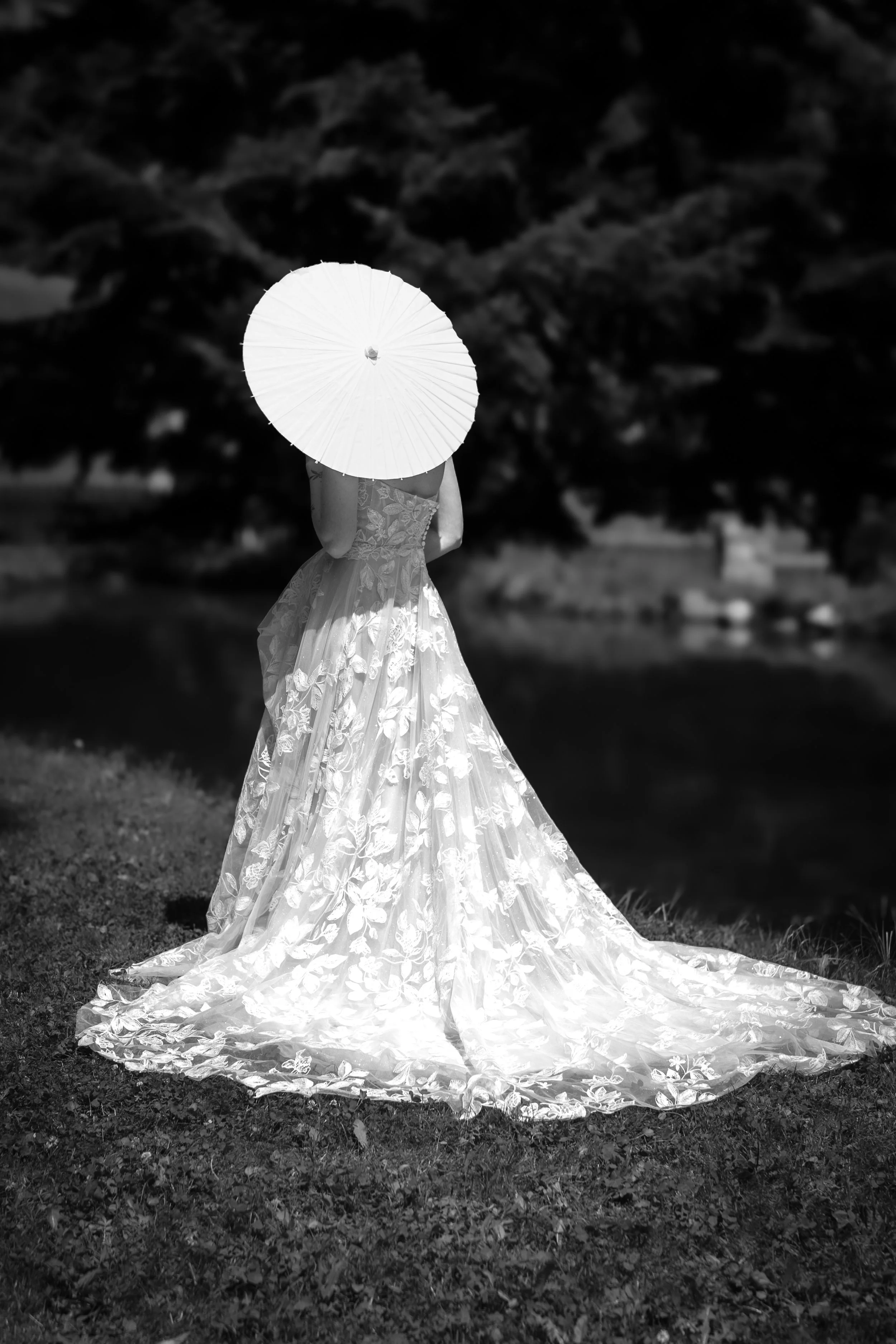 Eine Frau in einem langen, eleganten, weißen Brautkleid mit floralem Muster steht im Freien auf grünem Gras, hält einen weißen Sonnenschirm vor das Gesicht und steht vor einem großen Baum im Hintergrund.