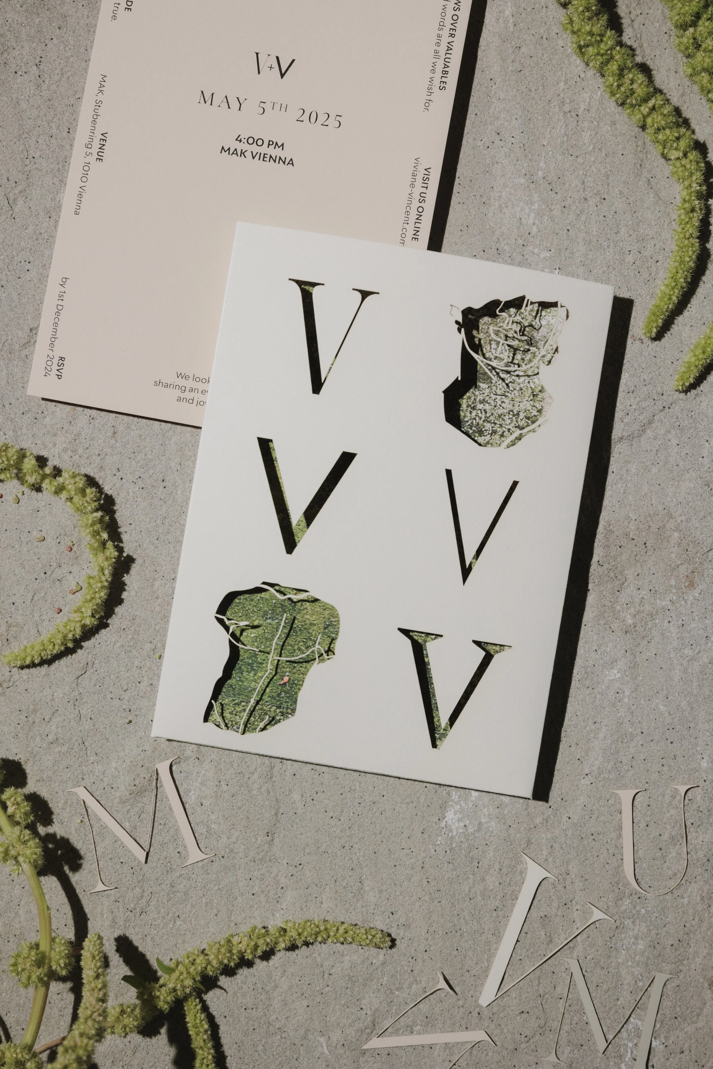 Ein Einladungskarten mit der Aufschrift 'V + V', Datum 5. Mai 2025, 16:00 Uhr, Ort: Mak Vienna, auf einem grauen Steintisch, umgeben von grünen Pflanzen.