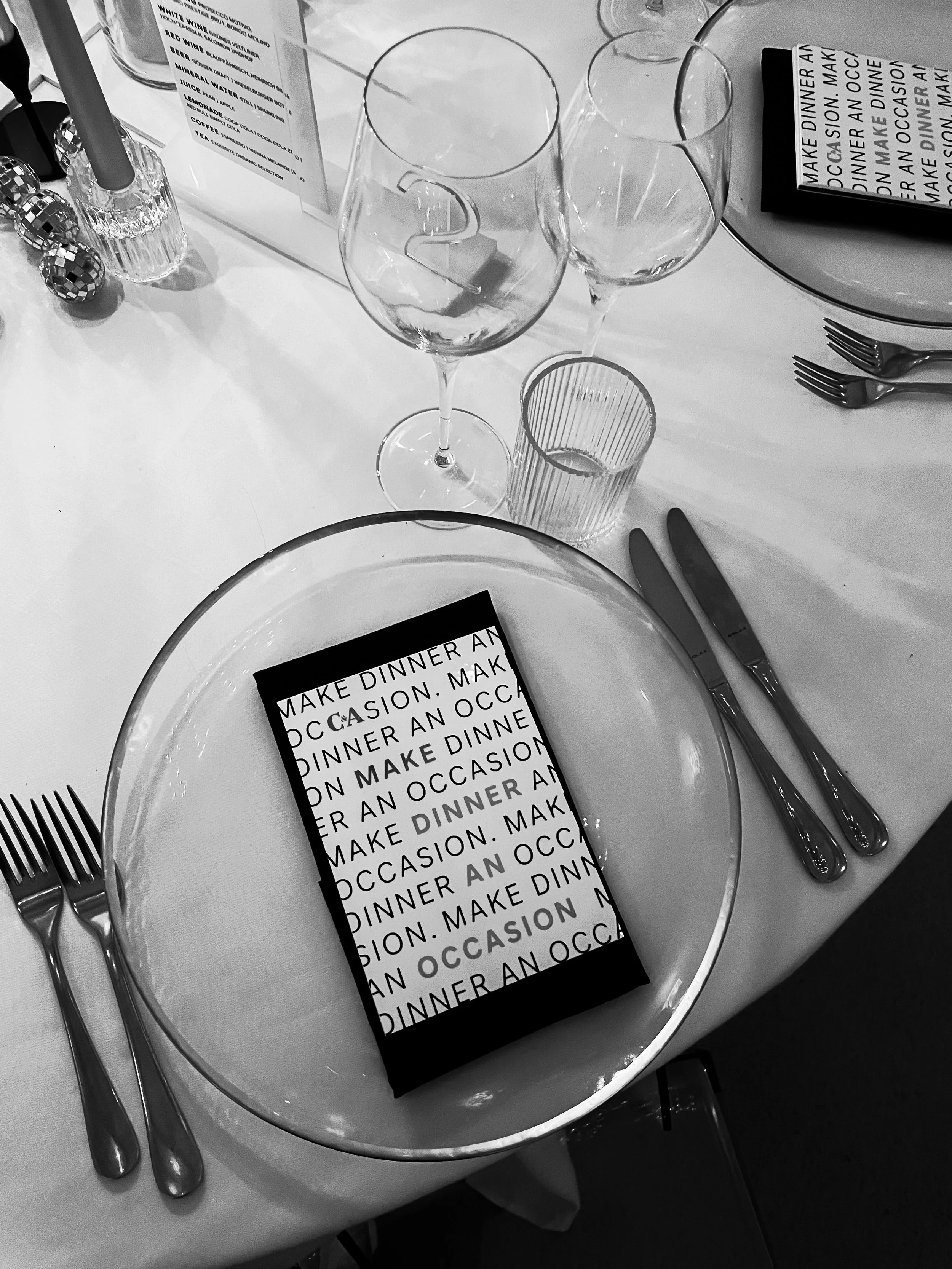 Ein festlich gedeckter Esstisch mit leeren Tellern, Besteck, Weingläsern, Wasserbechern, Kerzen und Partykugeln, dazu eine schwarze Einladungskarte mit weißer Schrift, auf der steht: 'MAKE DINNER AN OCCASION'