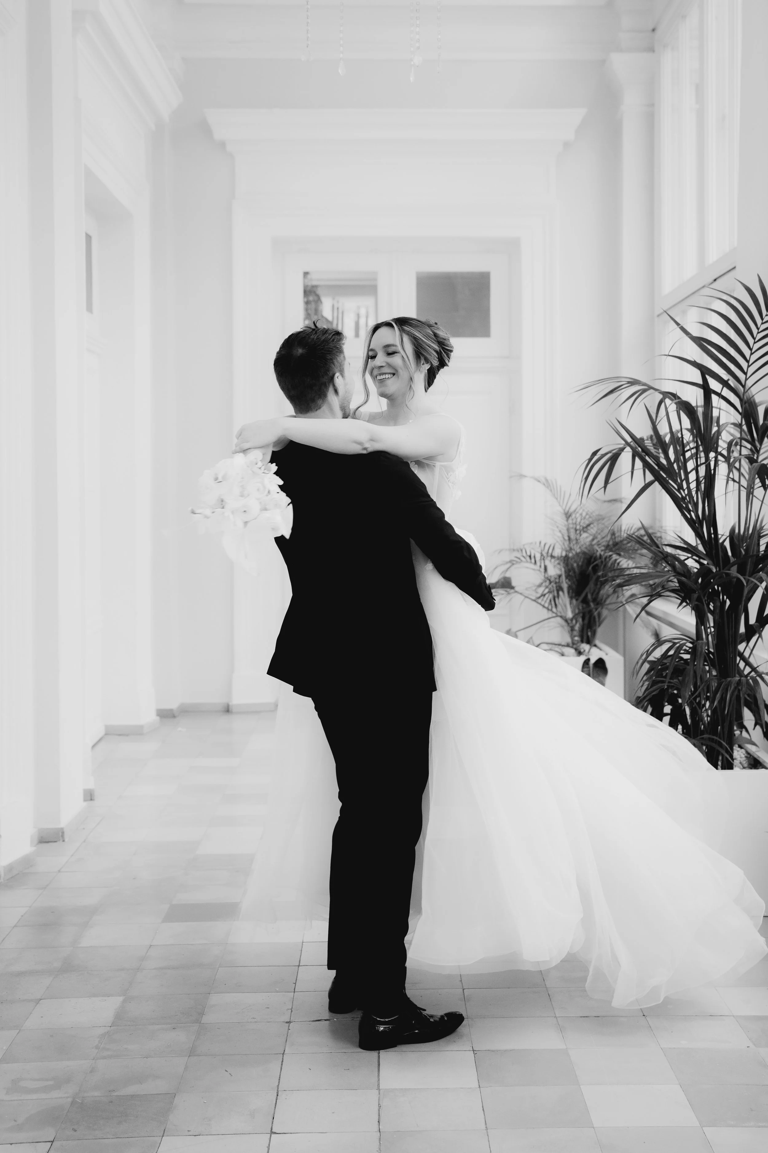 Braut und Bräutigam tanzen in einem hellen Flur, die Braut lächelt, während sie vom Bräutigam gehalten wird, im Schwarz-Weiß-Foto.