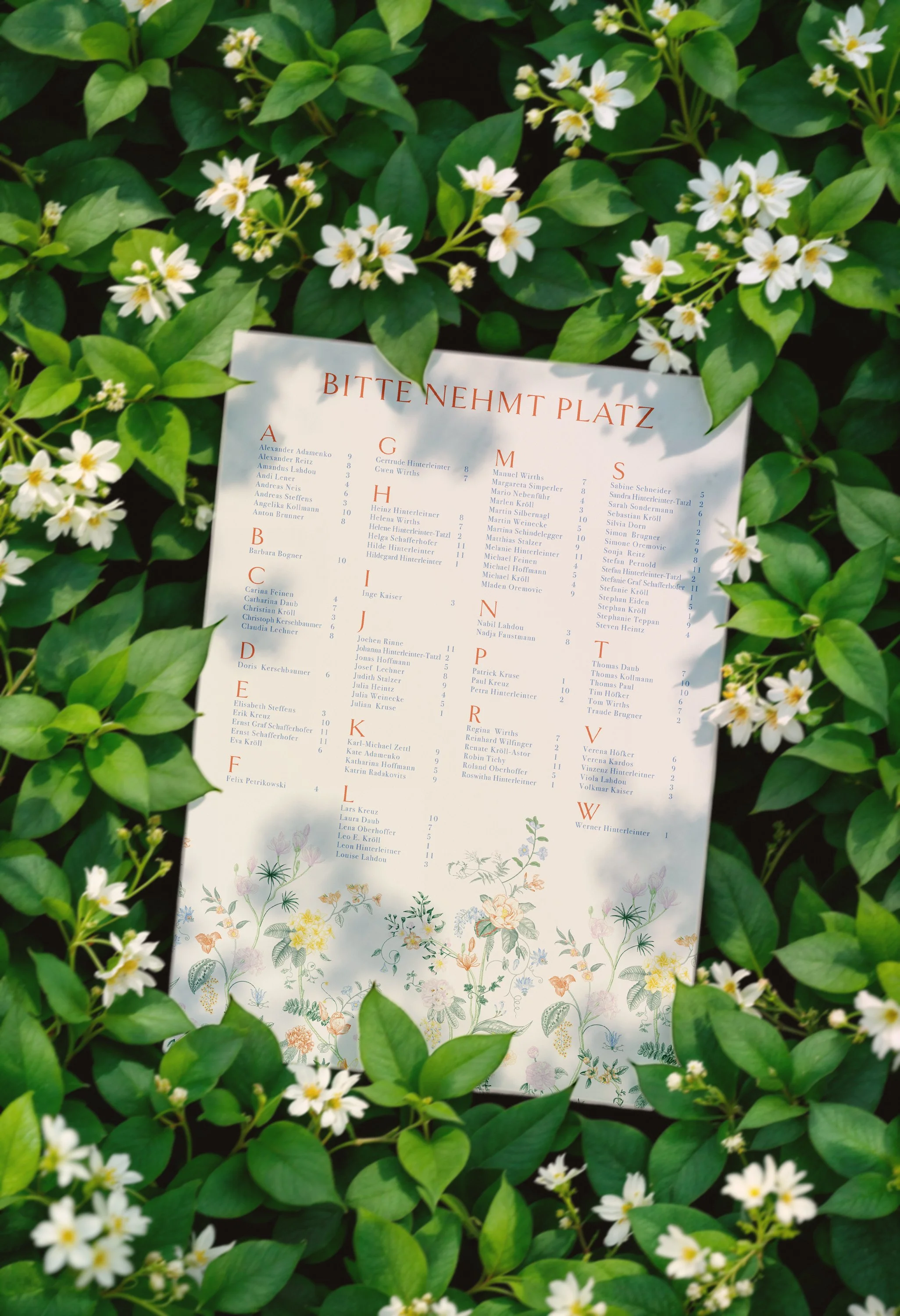 Ein elegantes Sitzplanschild A1 'BITTE NEHMT PLATZ' liegt auf grünem Laub und ist mit Illustrationen von bunten edlen Blumen gestaltet.