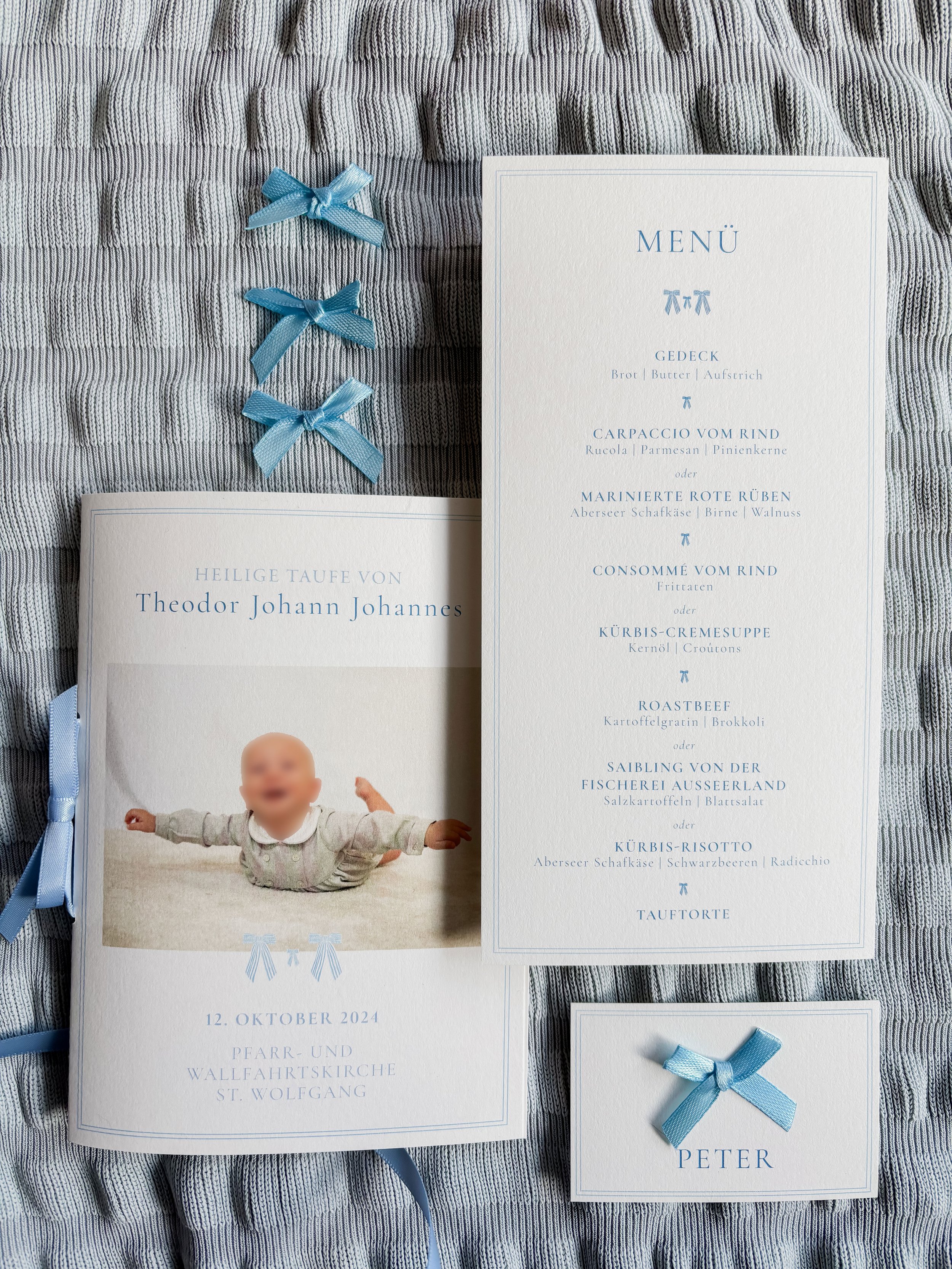 Ein Pinkleinenalbum mit einem Babyfoto, einen Menü-Plan für eine Taufe, blauen Bändern und einem Namensschild mit dem Namen Peter auf einem gestreiften Stoff.