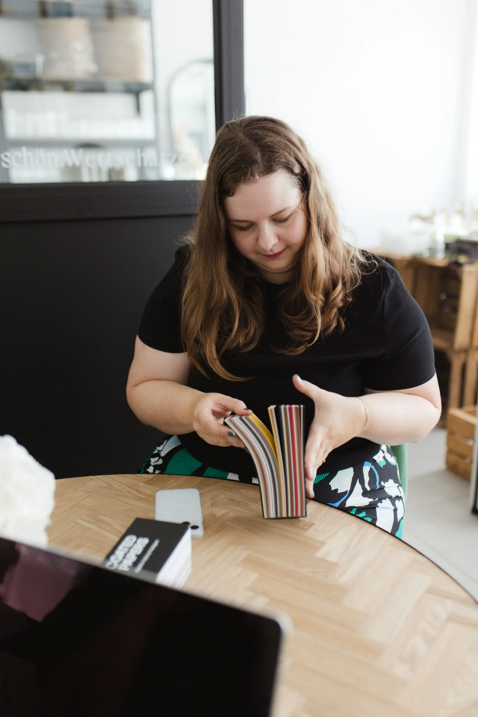 Eine Frau mit langen, lockigen, braunen Haaren sitzt an einem runden Holztisch, betrachtet ein offen liegendes Buch mit bunten Streifen im Inneren, in einem modernen Raum mit großen Fenstern und dunkler Wand im Hintergrund.