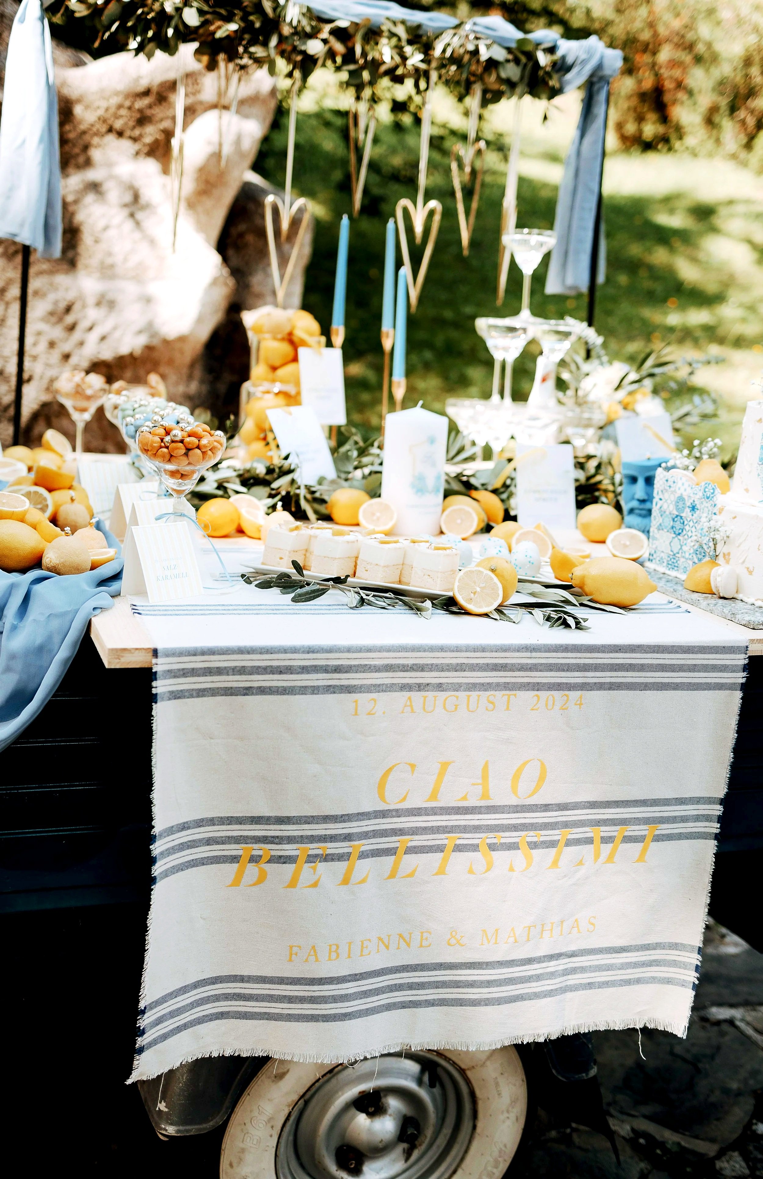 Ein festlich gedeckter Tisch auf einem Wagen mit einer Tafel, die den Namen CHO BELLISSIMA und das Datum 12. August 2024 trägt. Der Tisch ist mit Zitronen, Kerzen, Kuchen und Dekorationen in Blau- und Gelbtönen geschmückt.