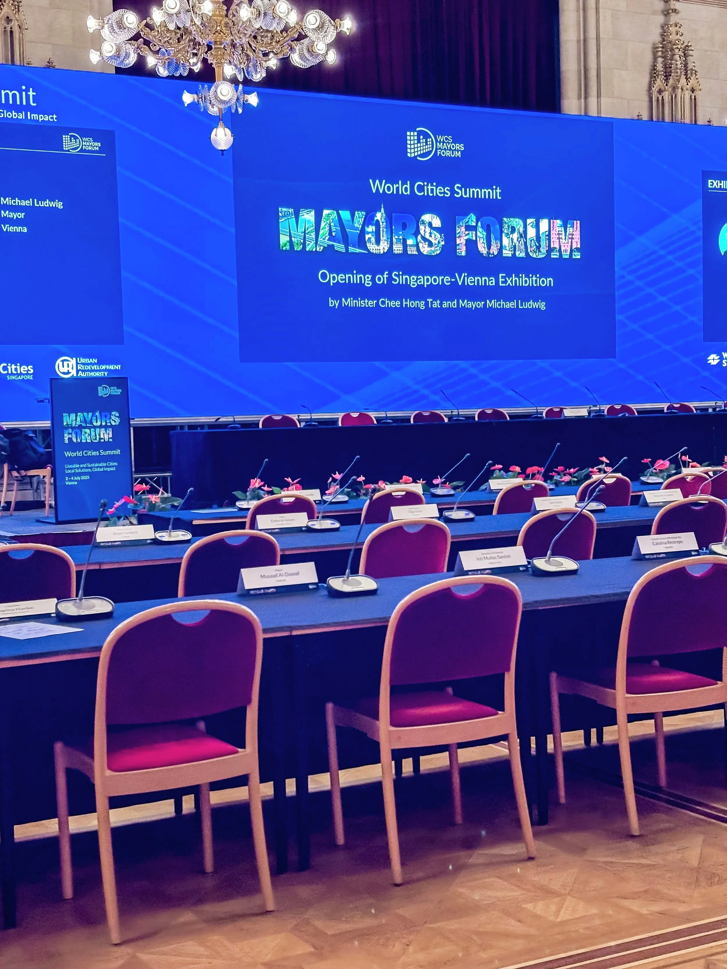 Leeres Konferenzzimmer mit Tischen, roten Stühlen, Mikrophonen und Namensschildern vor einer großen digitalen Anzeige für den World Cities Summit Event. Der Bildschirm zeigt den Titel "World Cities Summit MAYORS FORUM" und Hinweise auf die Eröffnung der Singapore-Vienna-Ausstellung.
