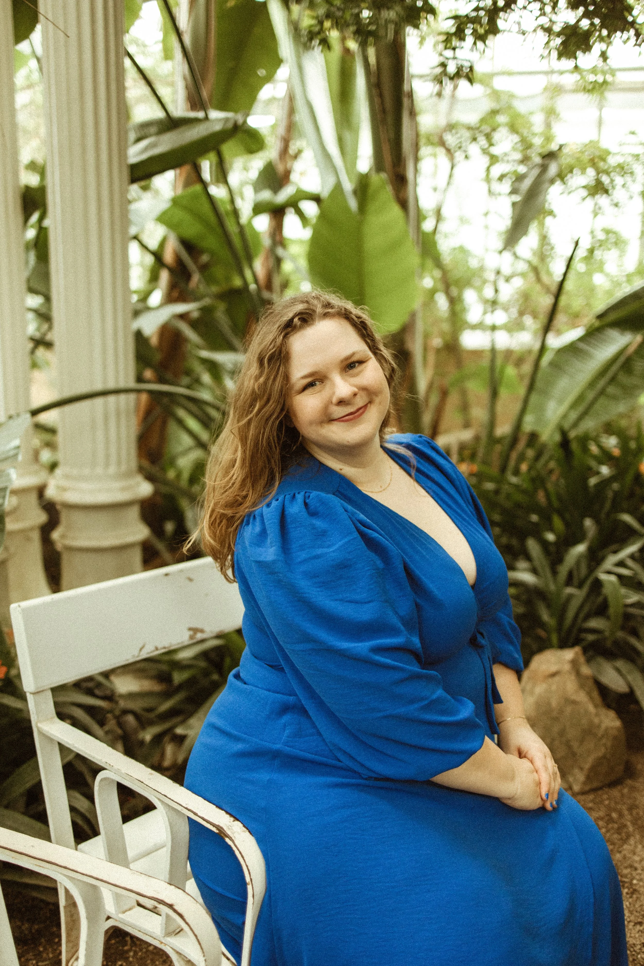 Eine lächelnde Frau in einem blauen Kleid sitzt auf einer weißen Bank in einer tropischen Pflanzenumgebung, umgeben von großen grünen Blättern und Pflanzen.