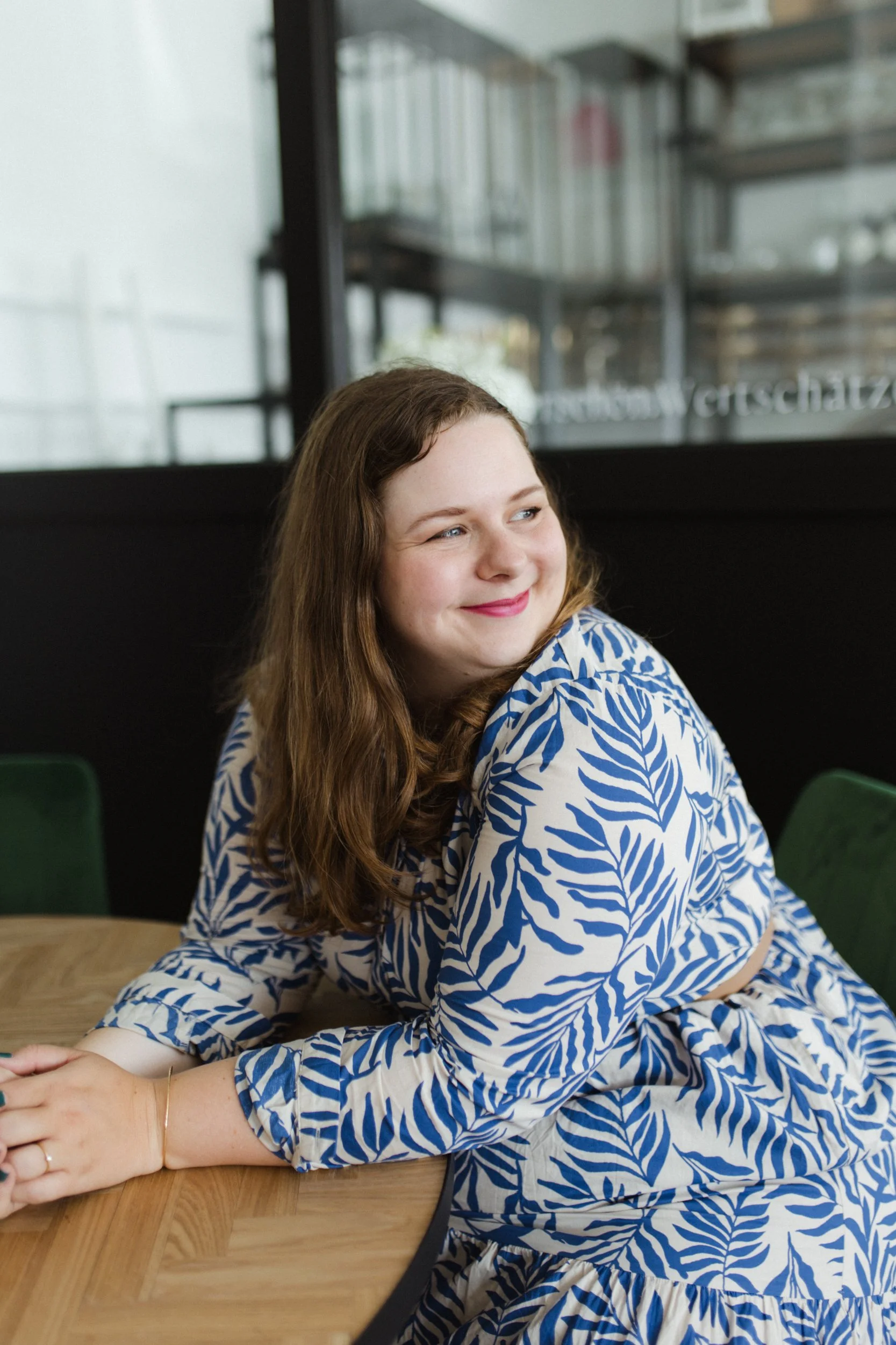 Ein Mädchen mit braunen Haaren, das auf einem Holztisch sitzt, trägt ein weiß-blau gemustertes Kleid mit Blättern. Sie lächelt und schaut zur Seite, im Hintergrund sind Regale und eine schwarze Wand sichtbar.