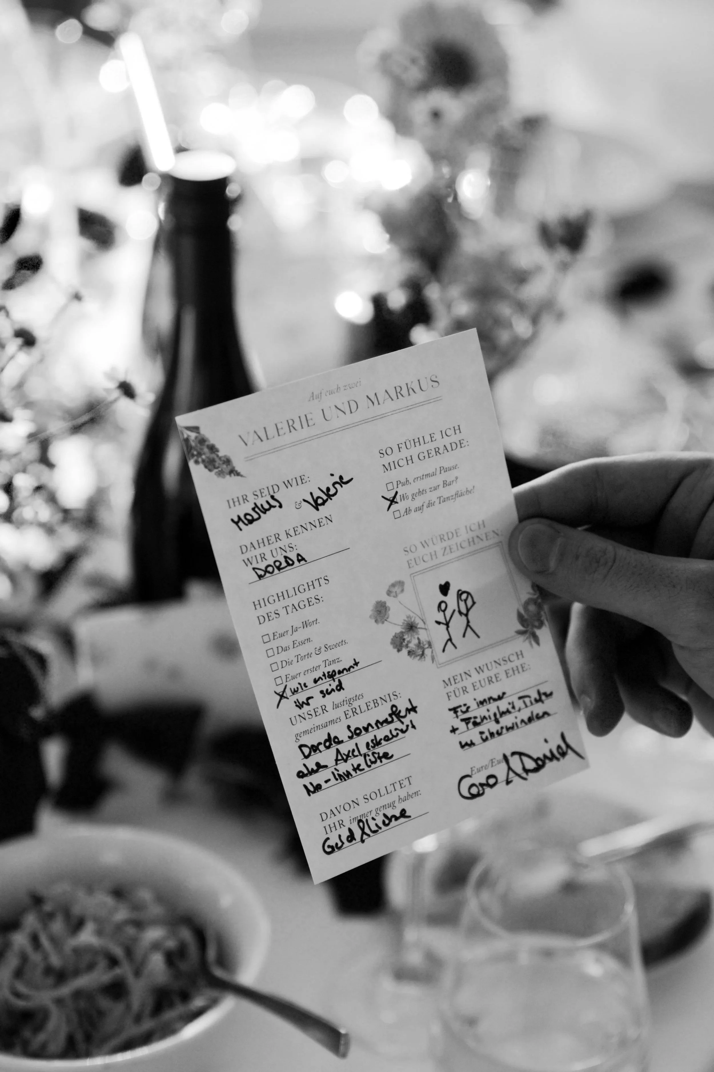 Eine Hand hält eine Sitzkarte bei einer Feier. Im Hintergrund sind eine Flasche, Gläser, eine Kerze und Blumen zu sehen, alles in Schwarz-Weiß.