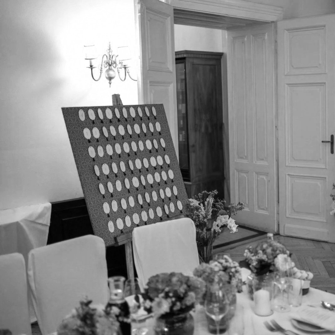 Ein festlich gedeckter Esstisch mit Blumen, Kerzen und Tellern in einem eleganten Raum mit Holzvertäfelung und einer Holzvitrine. Ein Wandbild oder eine Dekoration ist provisorisch auf einen Ständer gestellt.