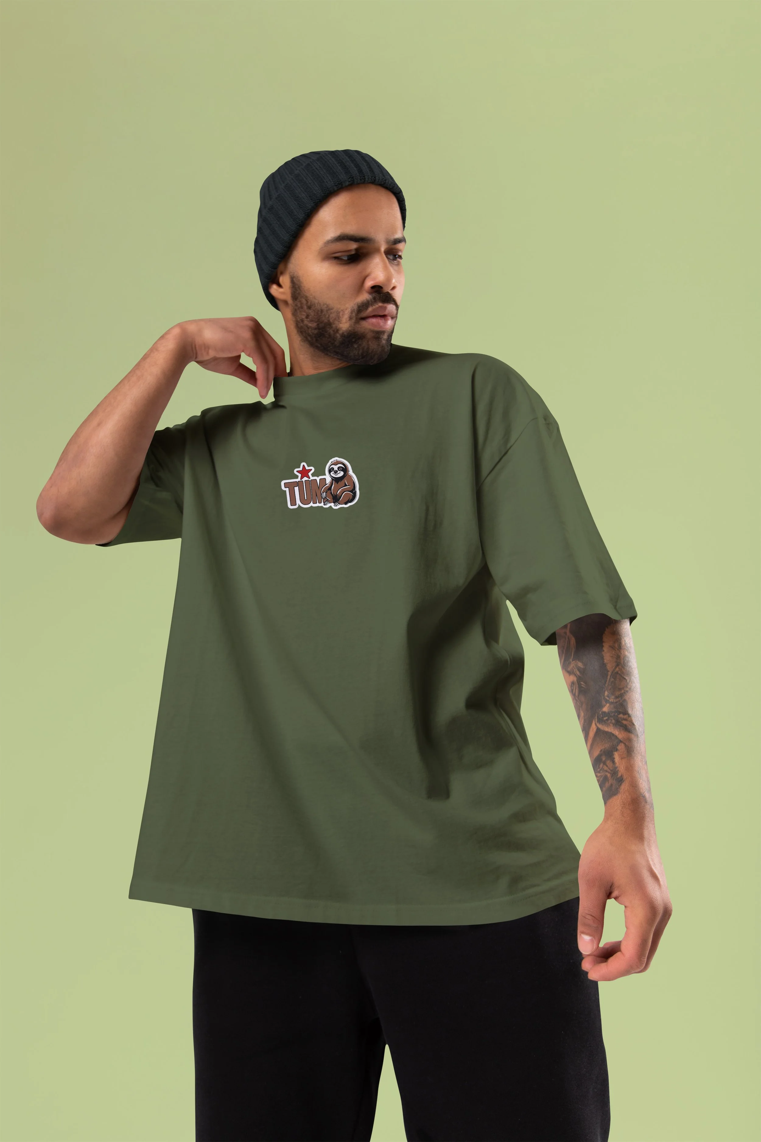 Model wearing olive green TUM T-shirt with sloth logo and black beanie, light green background.