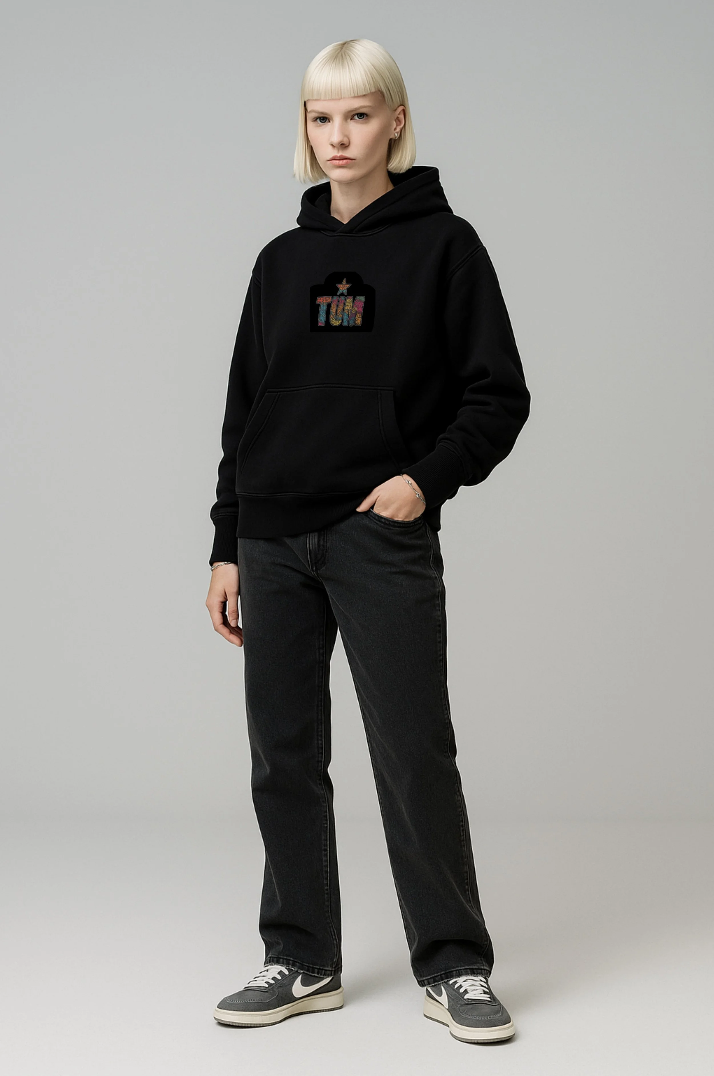 Young woman with blonde hair wearing a black hoodie with colorful text, black jeans, and gray Nike sneakers, standing against plain gray background.