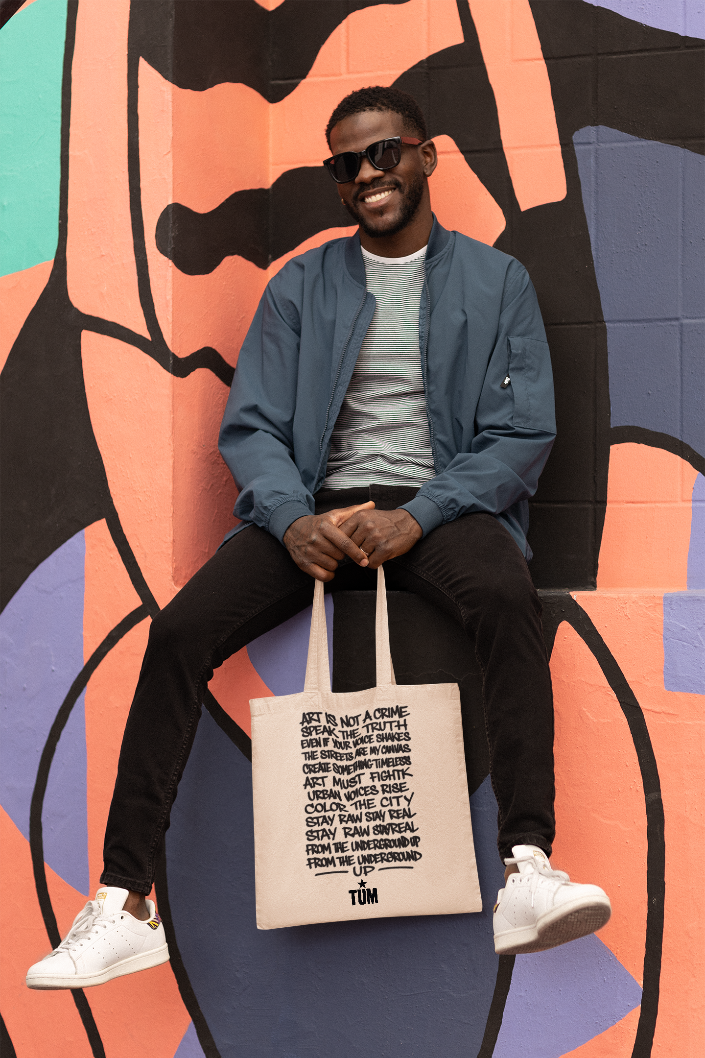 Model wearing blue jacket and sunglasses, sitting by graffiti mural with tote bag featuring handwritten text.