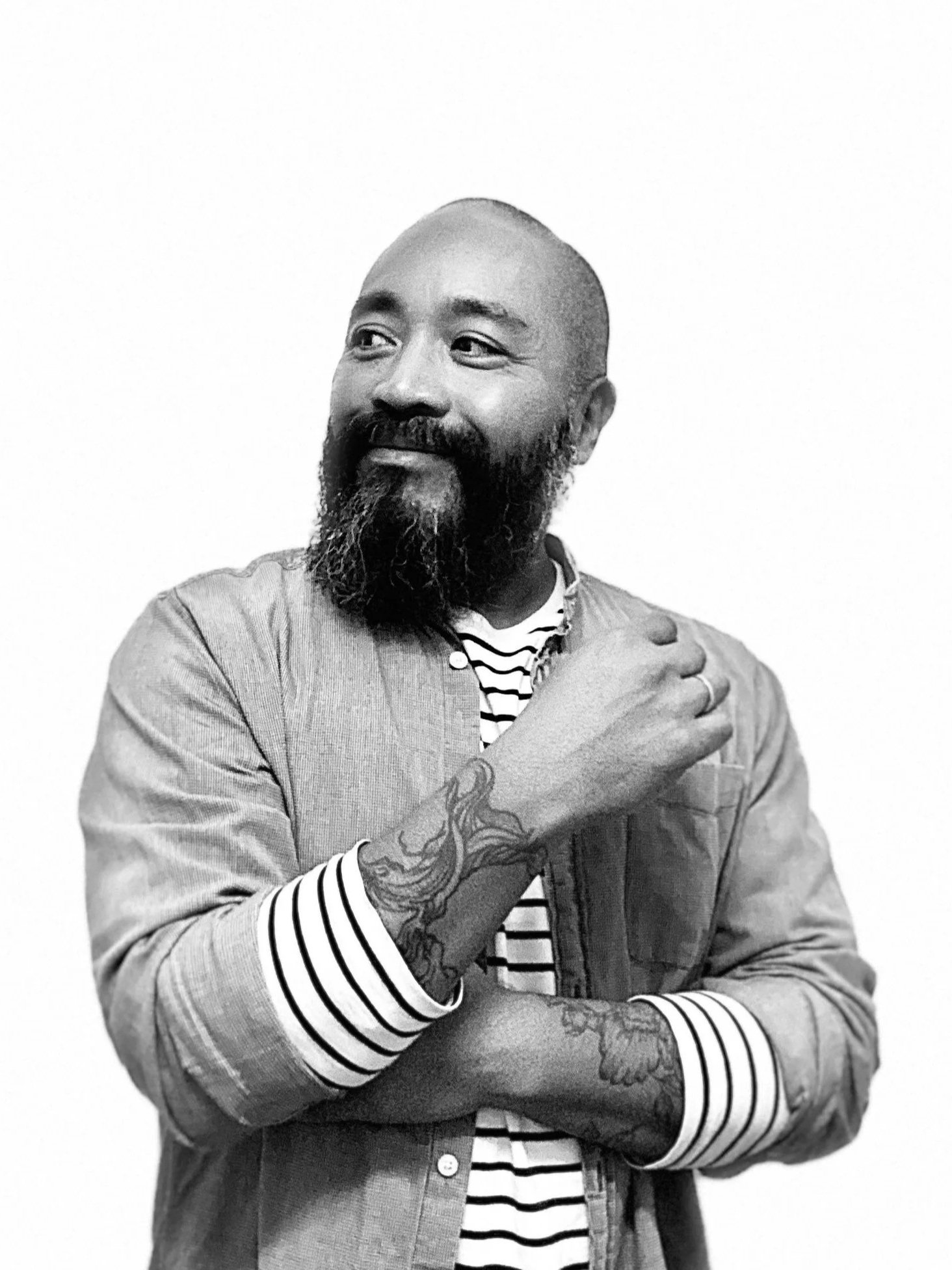 Black and white portrait of tattooed man in striped shirt and light jacket, looking to the side.