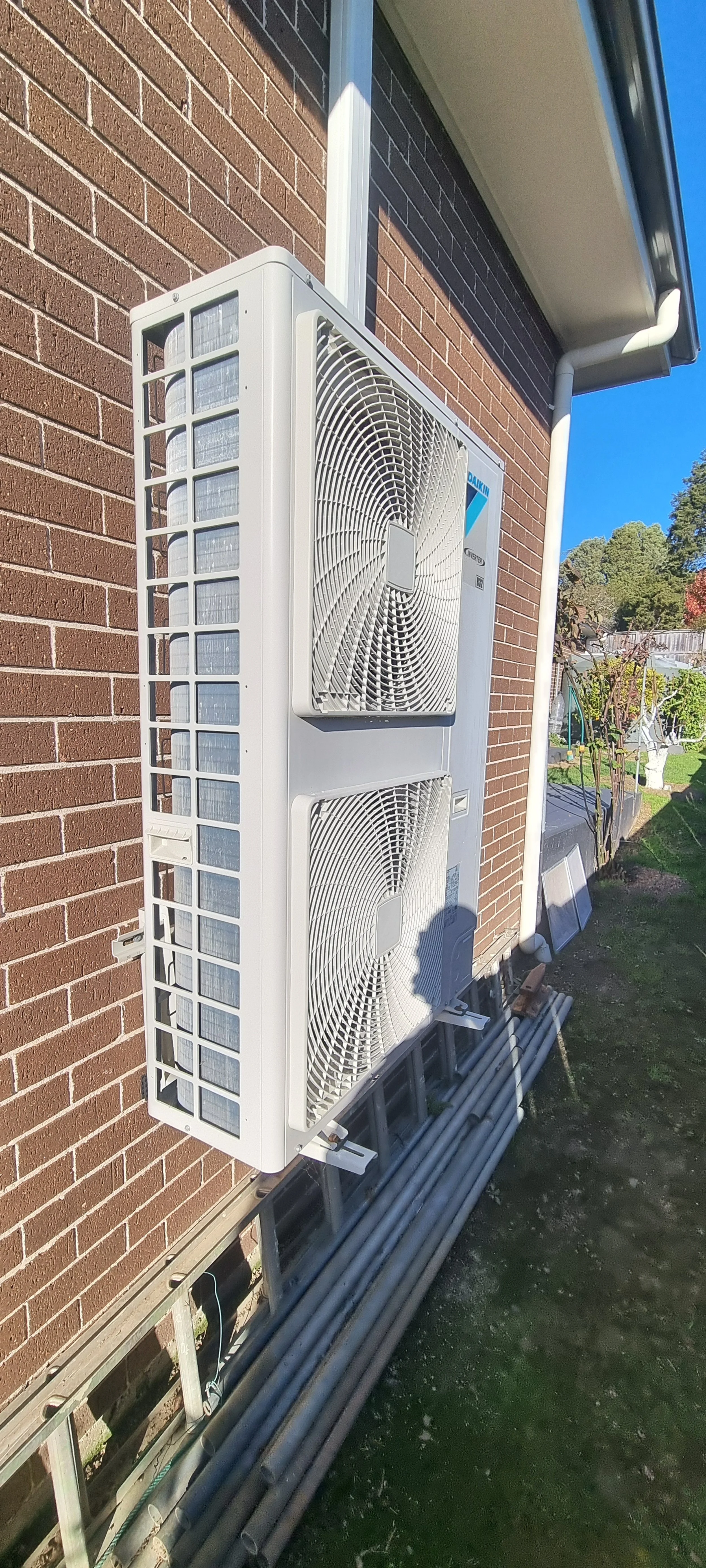 AC DC Southern Highlands Trade Services technician installation of outdoor air conditioning units mounted on a brick building wall, with pipes and a ladder beneath them, outdoors on a sunny day.