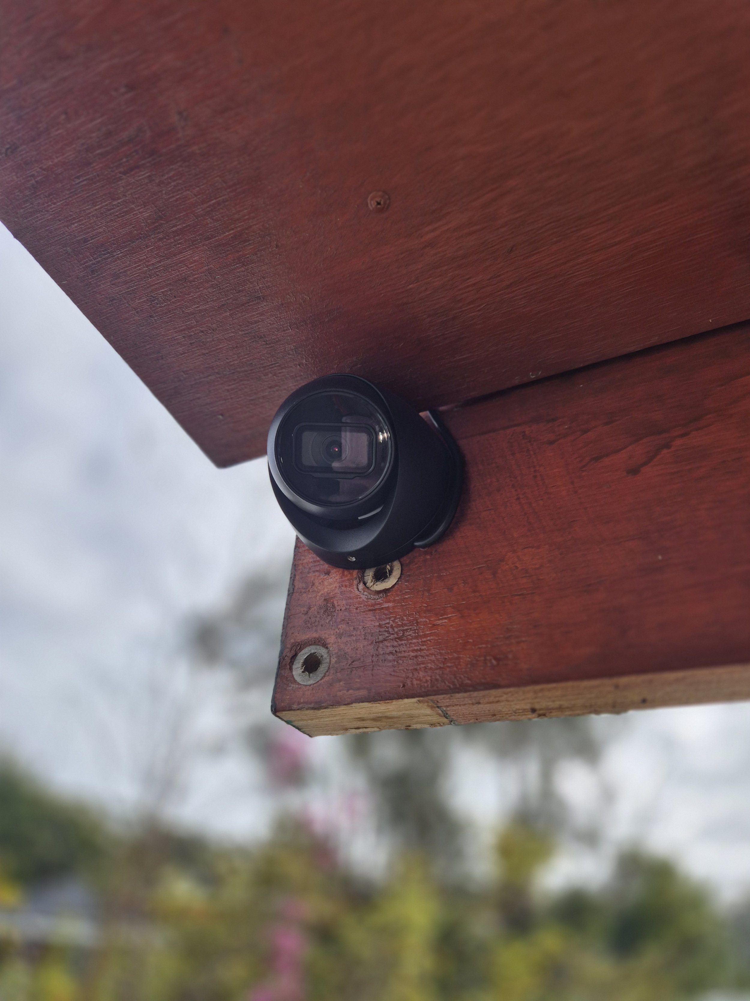 AC DC Southern Highlands Trade Services technician installed small black security camera mounted on a wooden outdoor surface.