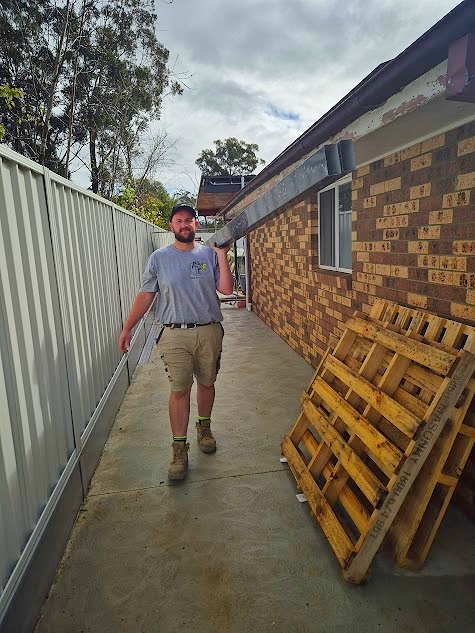 AC DC technician with a beard wearing a gray T-shirt, beige shorts, and work boots walking along a concrete pathway holding a long metal pole or pipe. To his right is a brick house with a window and a pile of wooden pallets leaning against the wall.