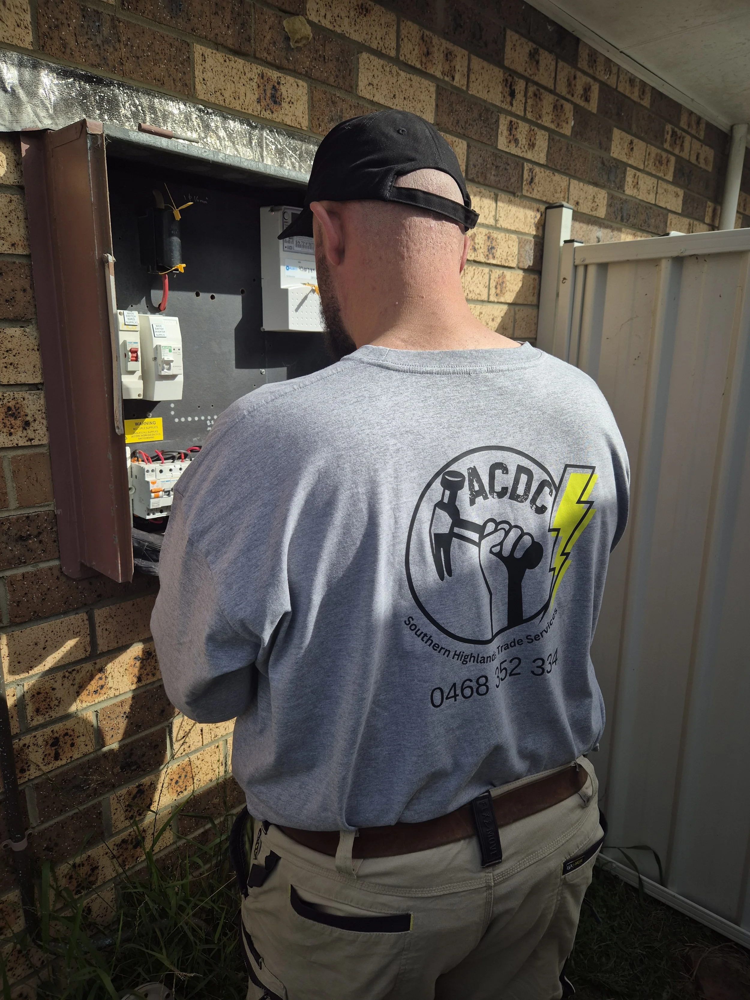 AC DC Southern Highlands Trade Services technician wearing a gray shirt and black cap working on an electrical box outside a brick wall.