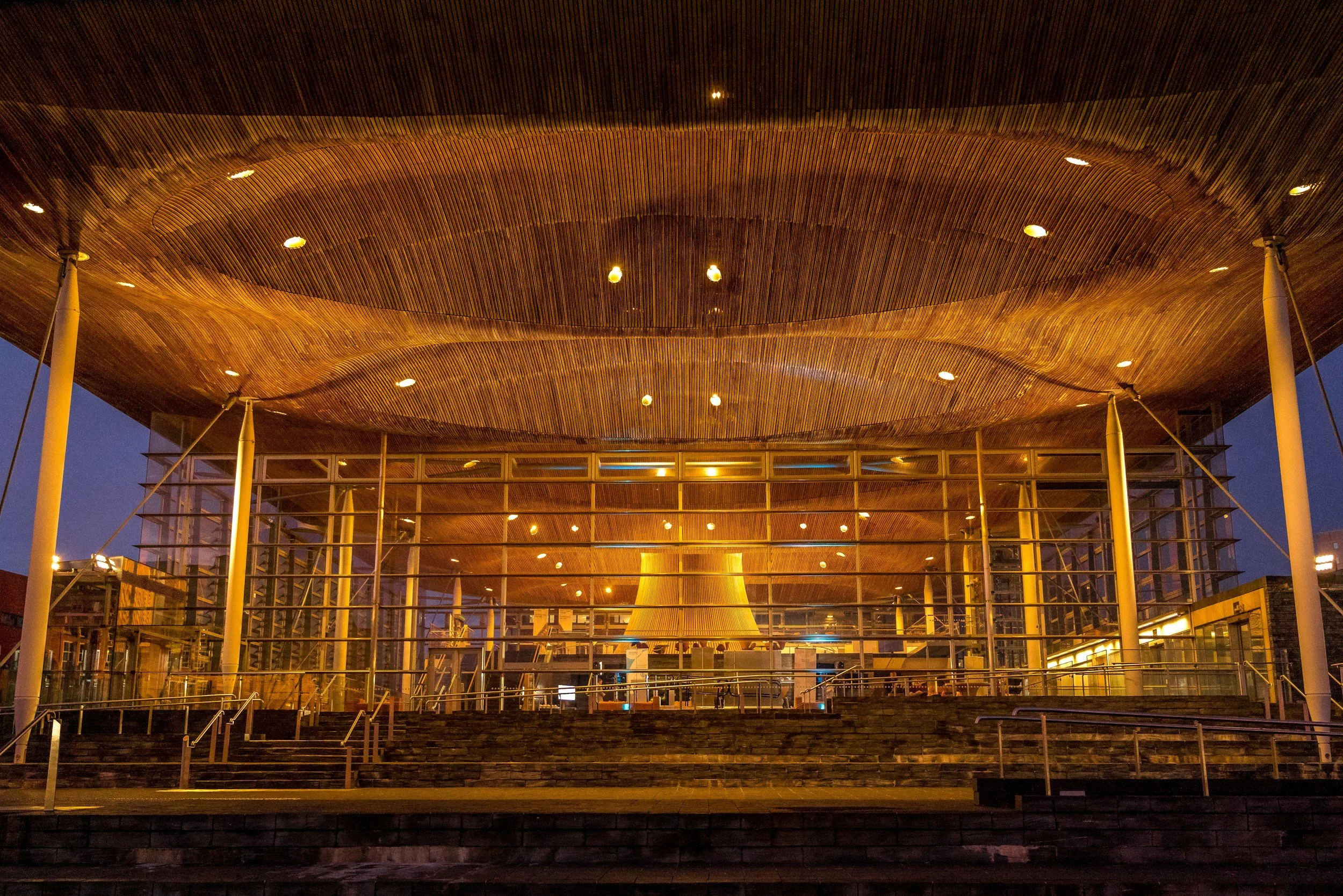 Night view of a modern building with a large, curved wooden roof, illuminated by yellow lights, with glass walls and surrounding stairs.