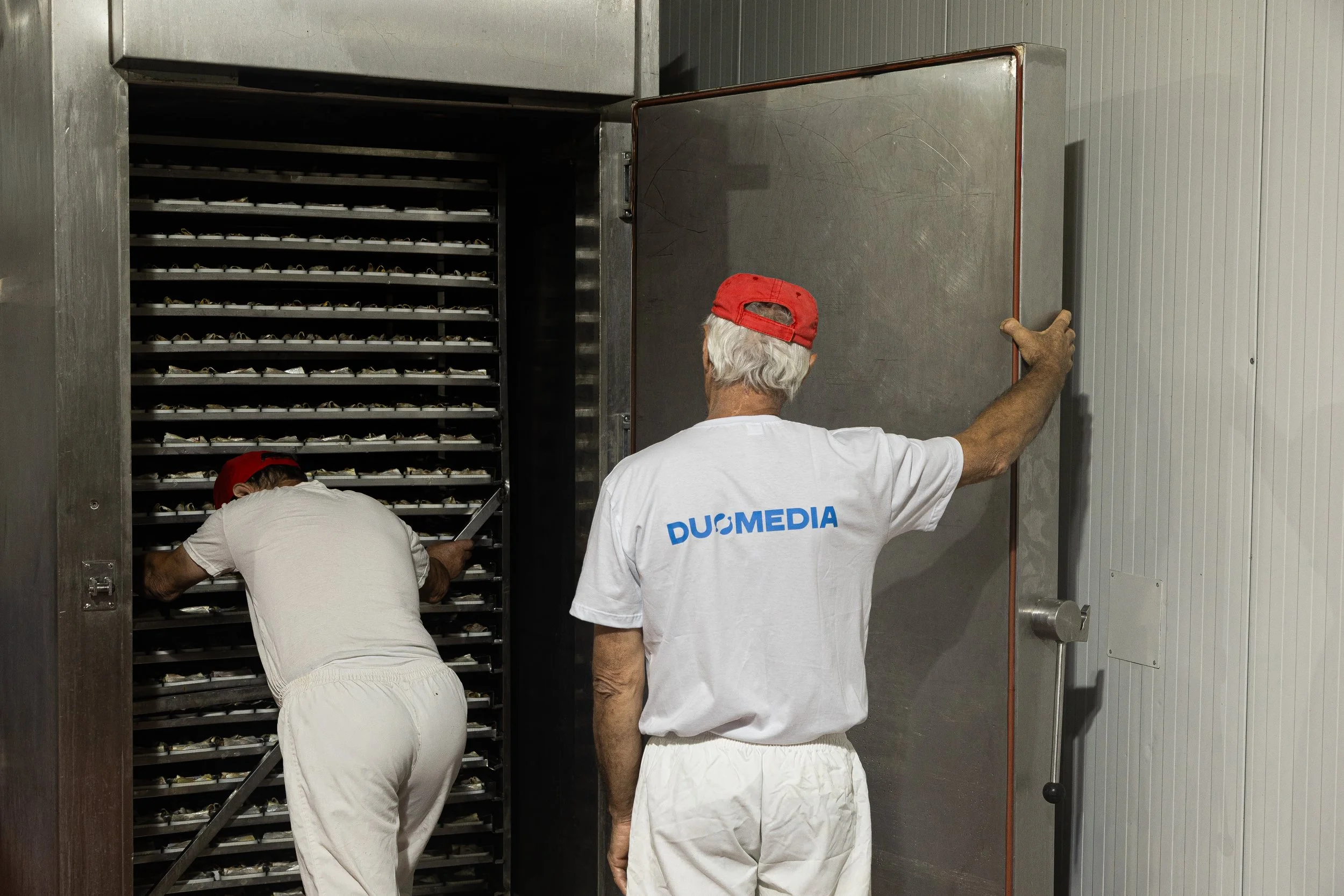 Two men working in a commercial environment, one opening a large metal door and the other examining small items on shelving inside a refrigerated or freezer room.