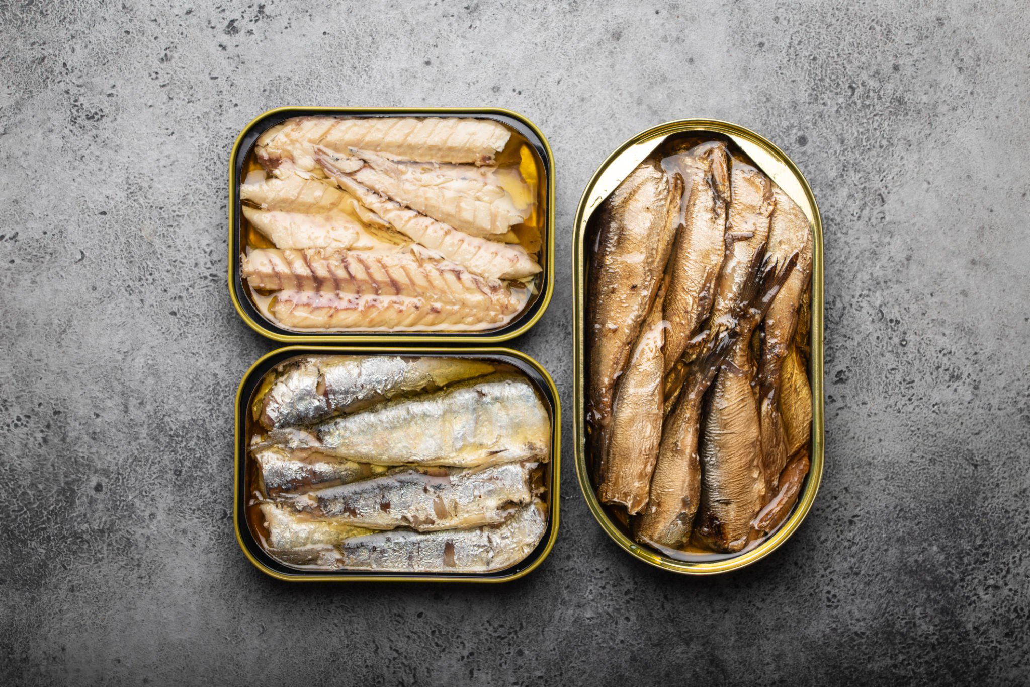 Three open tin cans filled with different types of preserved fish, placed on a gray stone surface.
