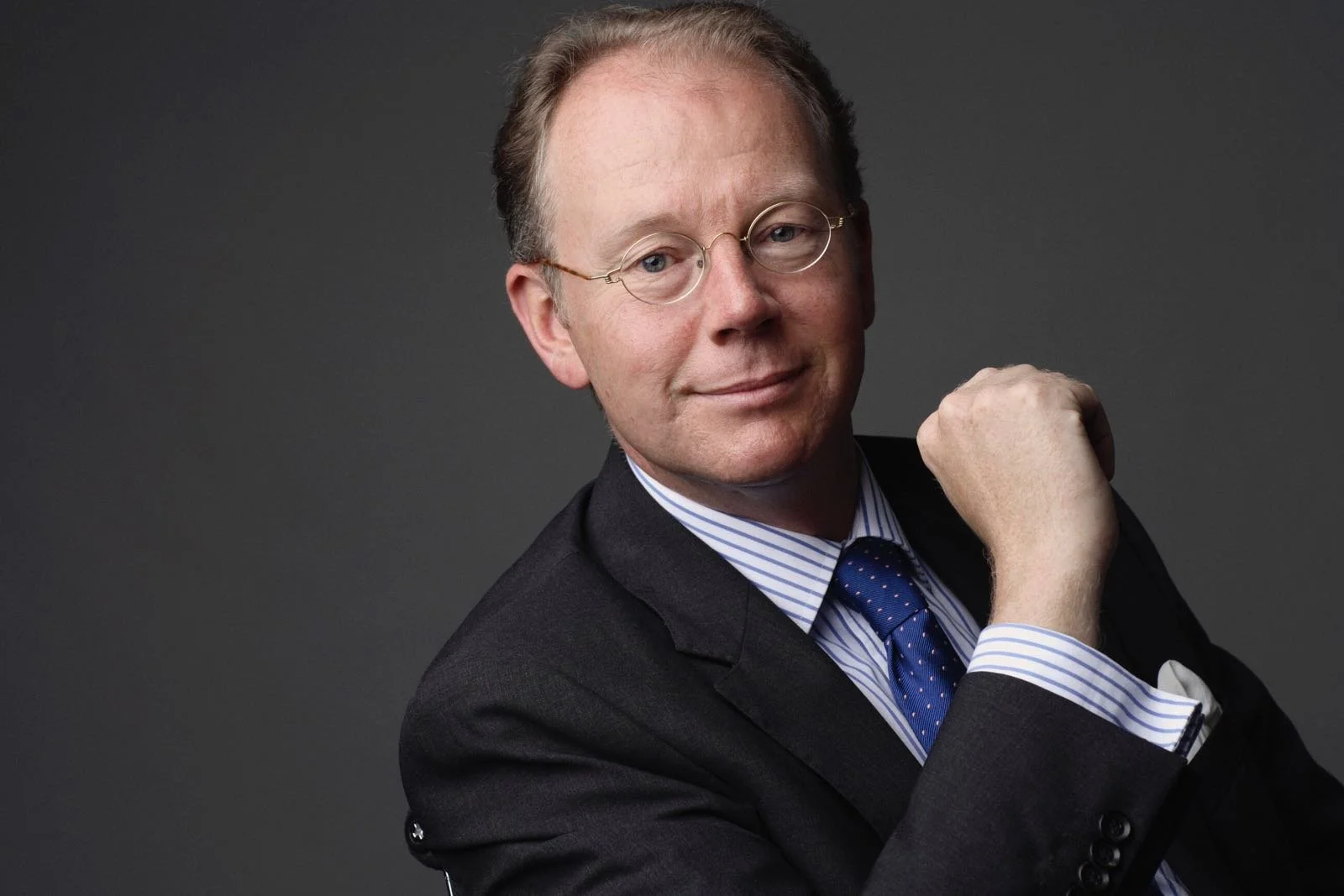 Man met bril, kostuum en stropdas, poseert met arm gebogen, glimlachend, tegen een donkere achtergrond