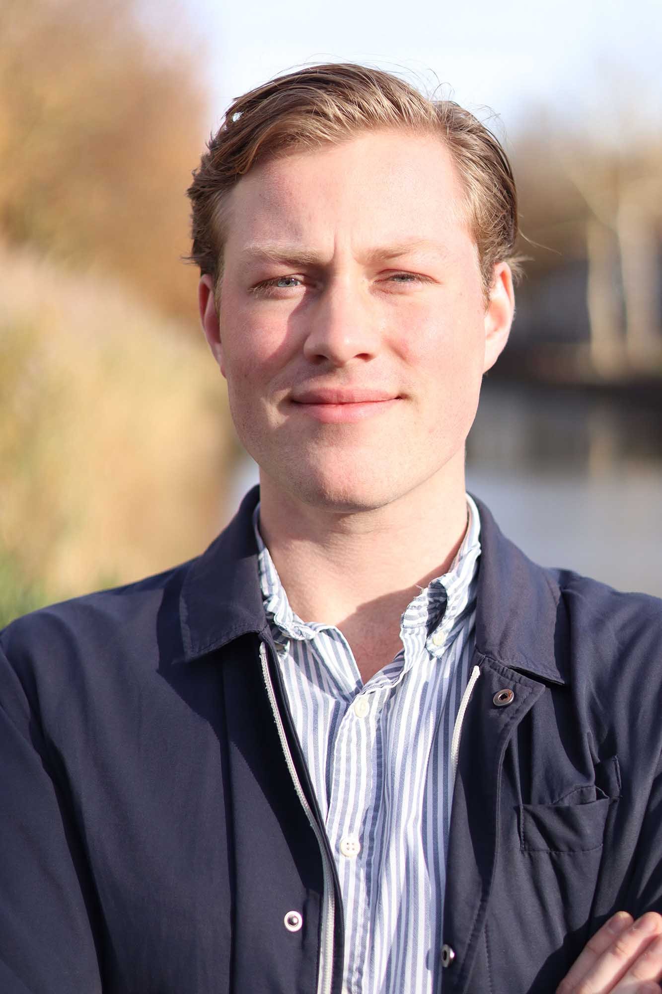 Portret van een jonge man met lichtbruin haar, gekleed in een blauwe jas en een gestreepte overhemd, standing outdoors bij zonlicht.